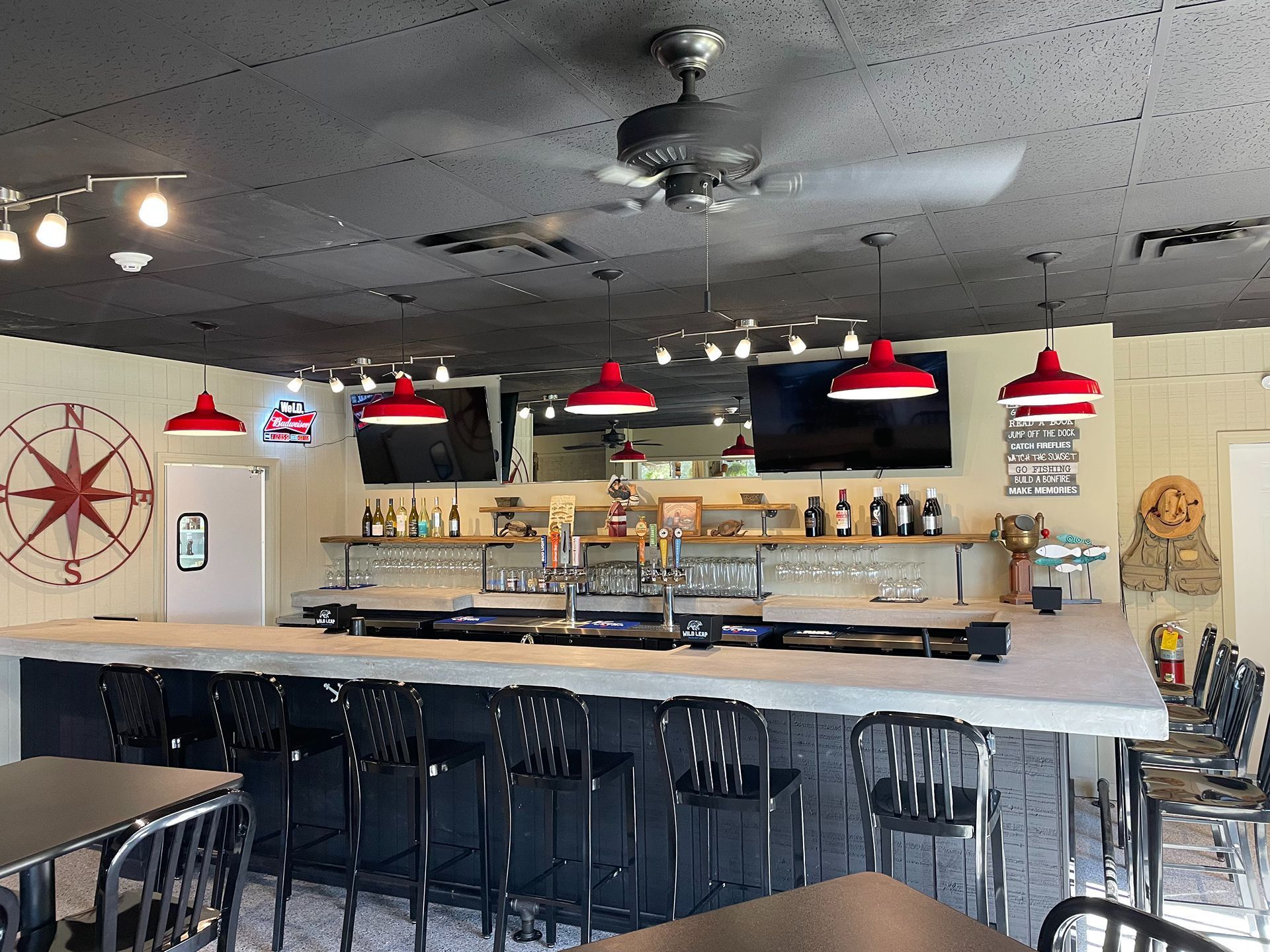 Bar interior with counter, stools, red pendant lights, compass decor, and televisions.
