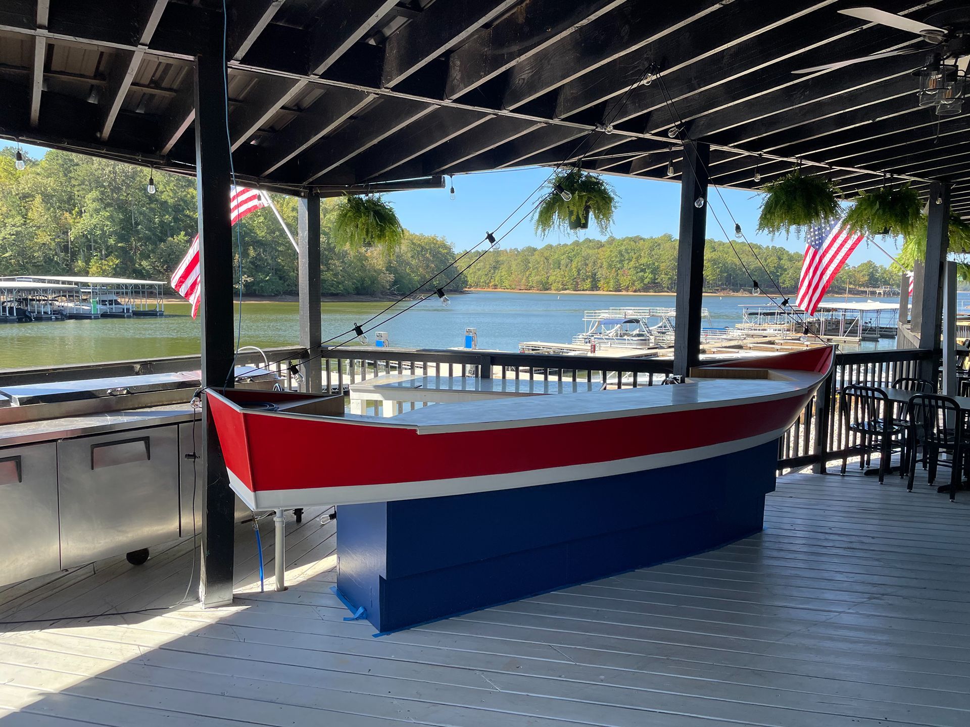 Boat-shaped outdoor bar overlooking a lake with blue sky. Red, white, and blue design with hanging plants.