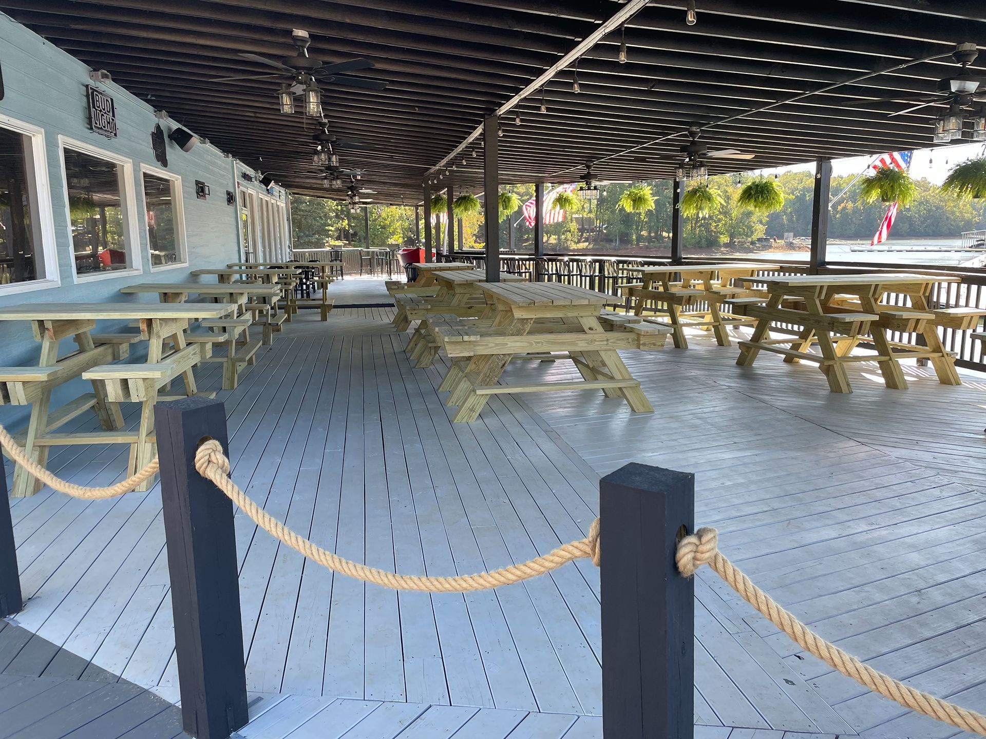 Outdoor dining area with picnic tables on a gray wooden deck under a black covered roof. Rope railing.