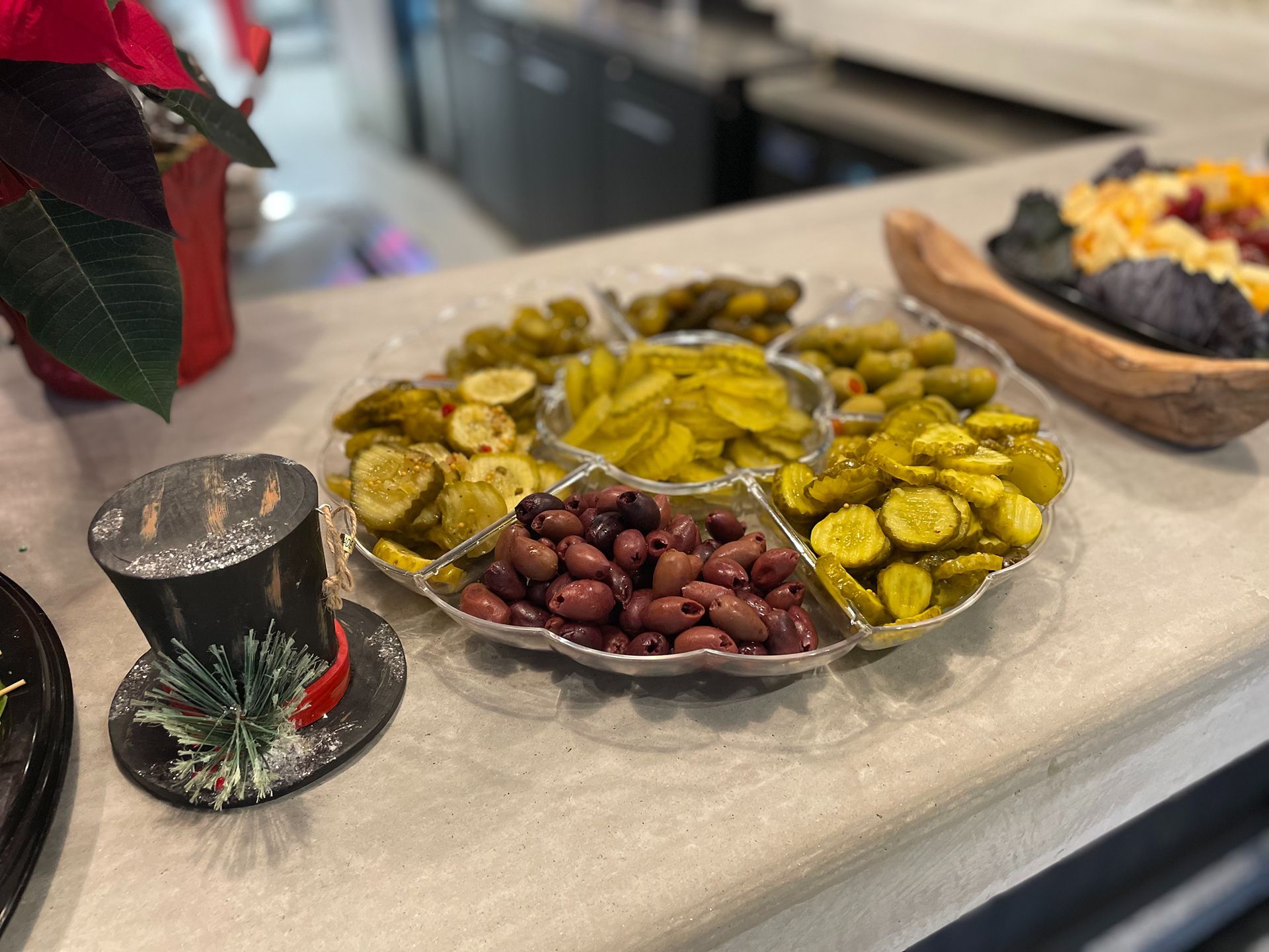 A clear platter with pickles and olives on a counter, with a decorative hat nearby.