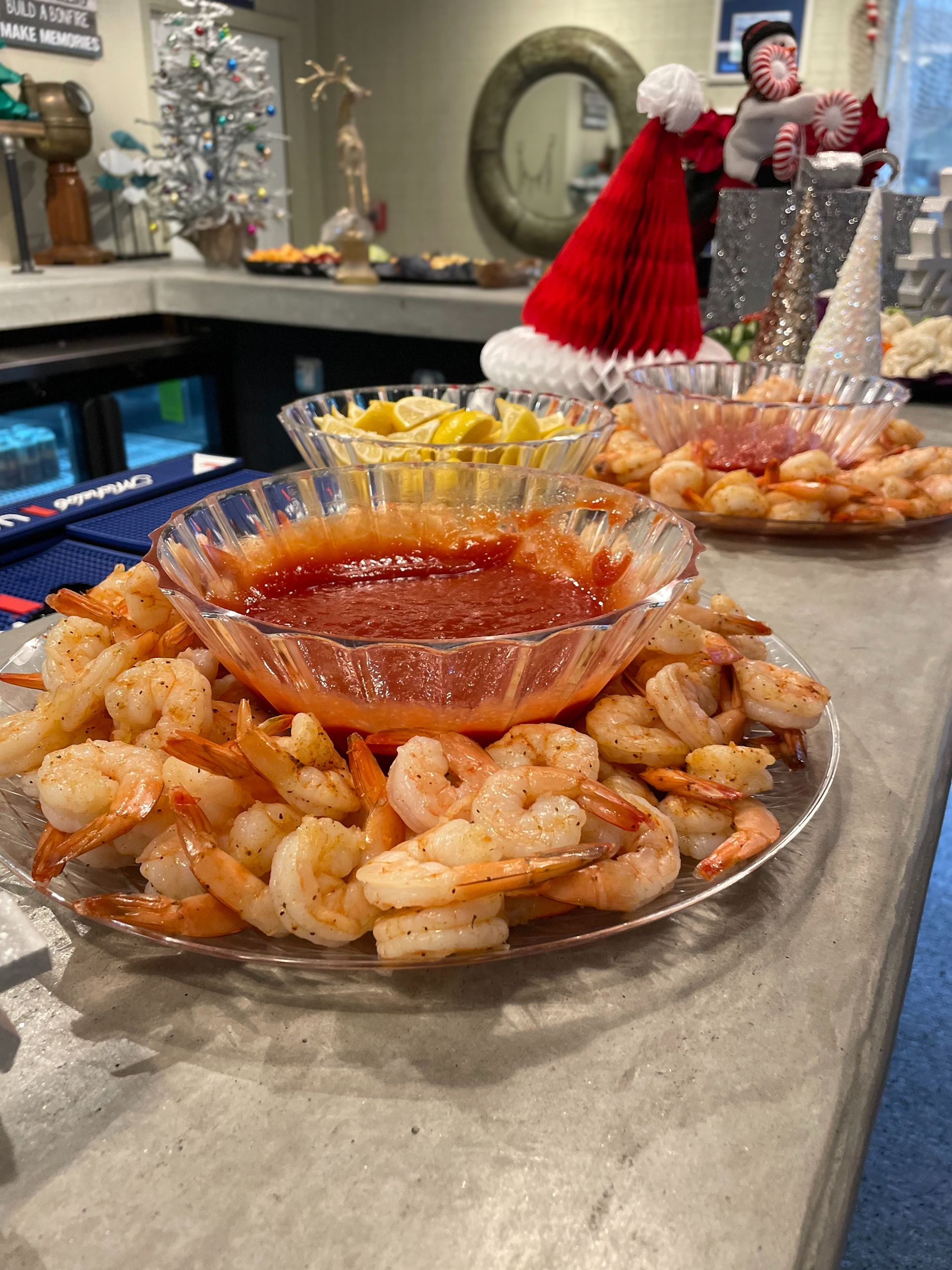 Shrimp cocktail platter with a bowl of red sauce, lemons, and Christmas decorations on a bar.