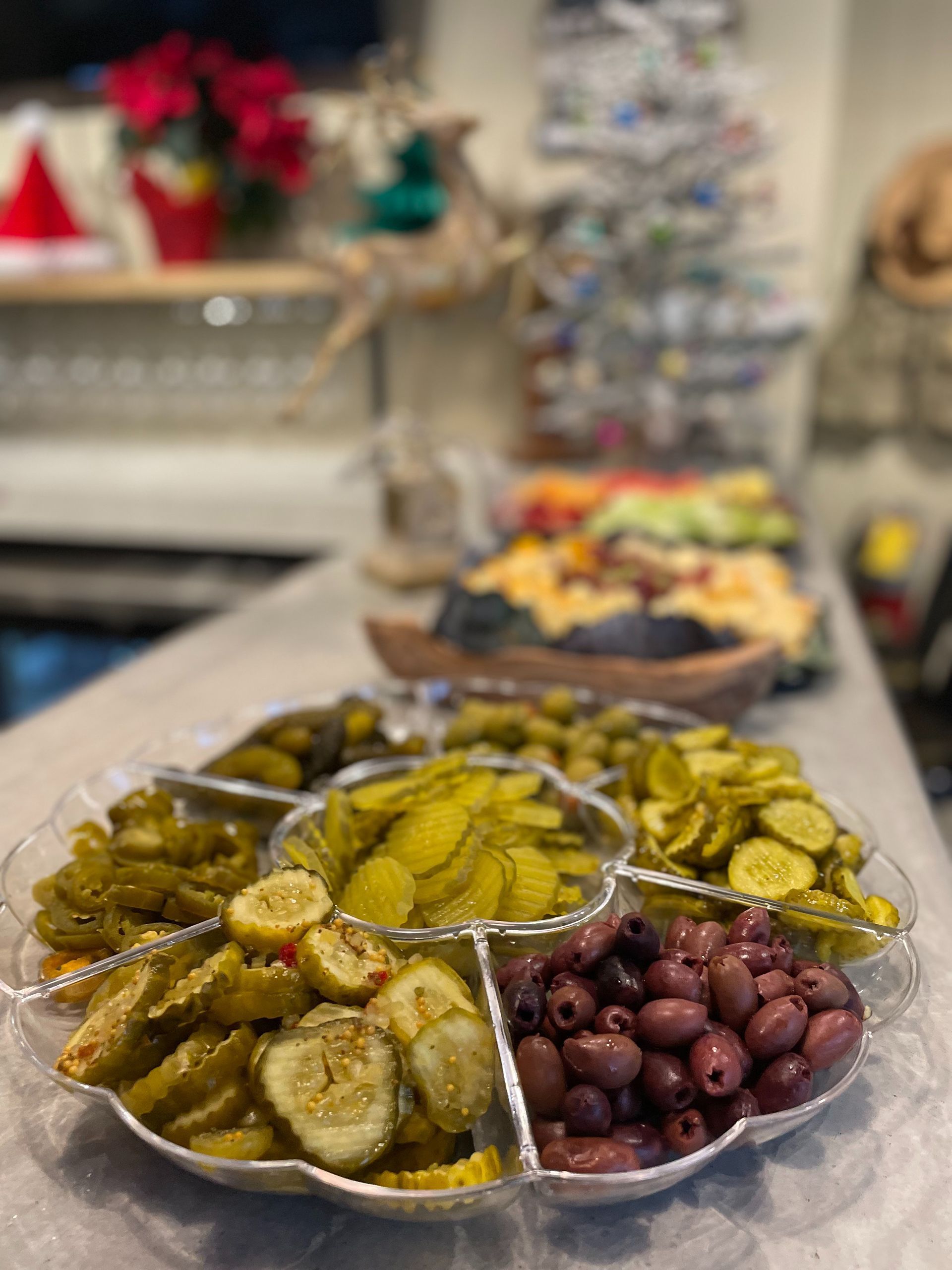 A platter of pickles and olives. Other snacks and a blurred Christmas tree are in the background.