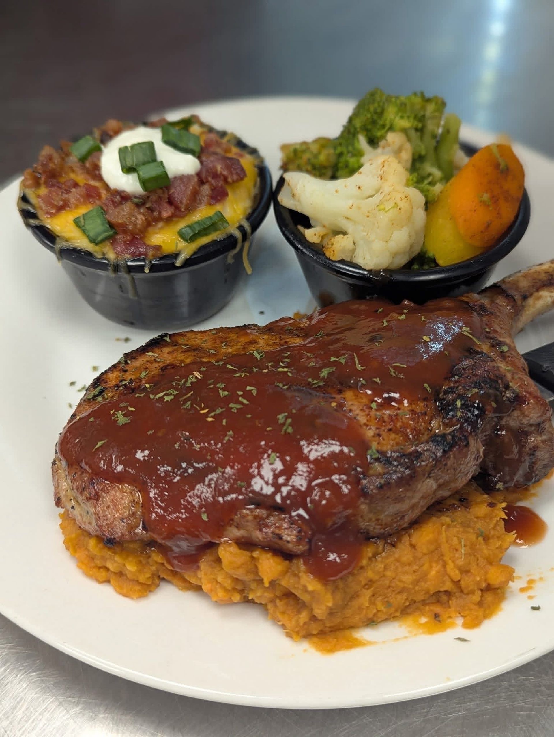 Plate of food: pork chop with sauce on sweet potato mash, side of loaded potato and mixed vegetables.