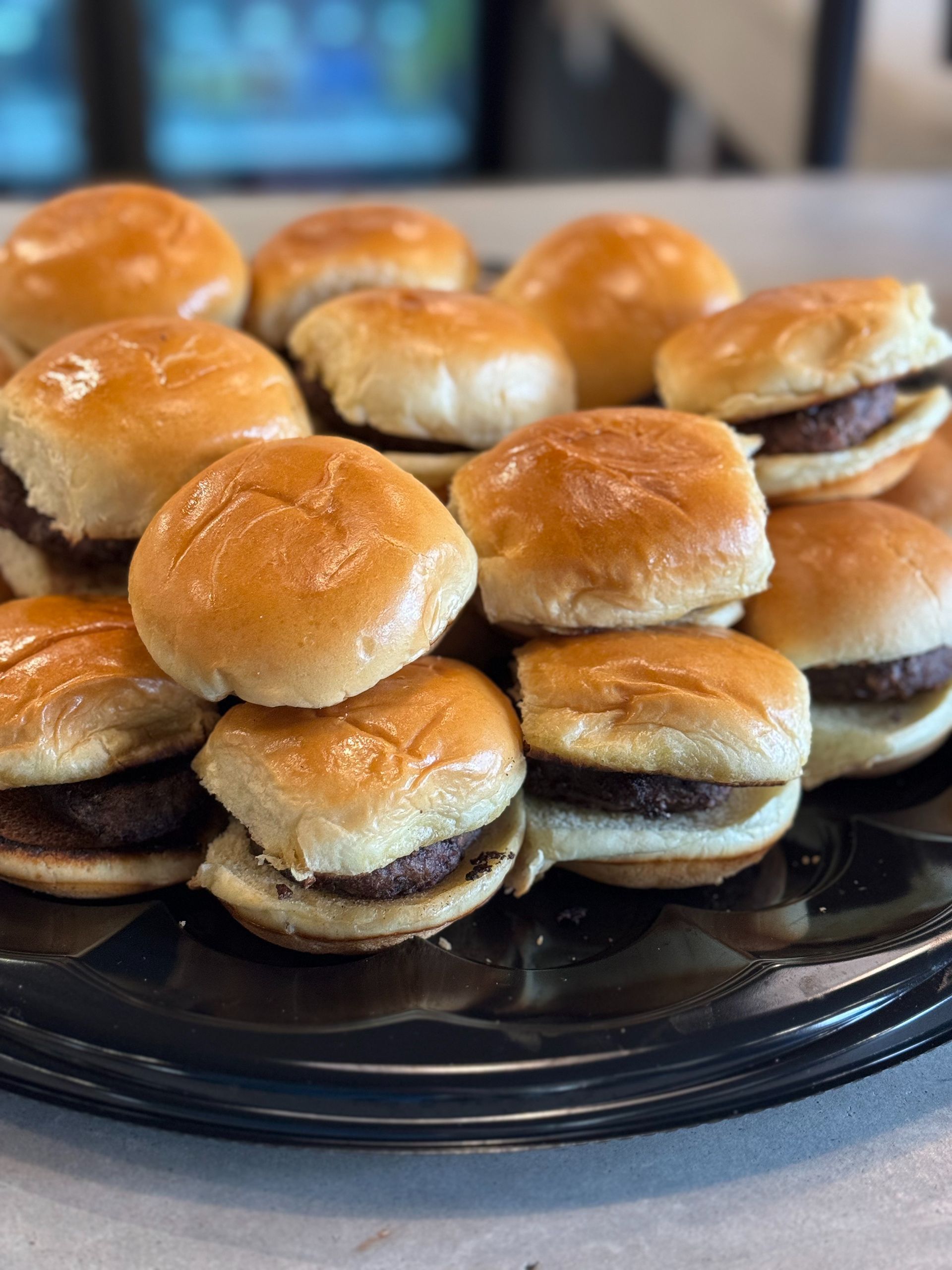 Miniature hamburgers on a black platter; several are stacked, showing golden buns and dark patties.