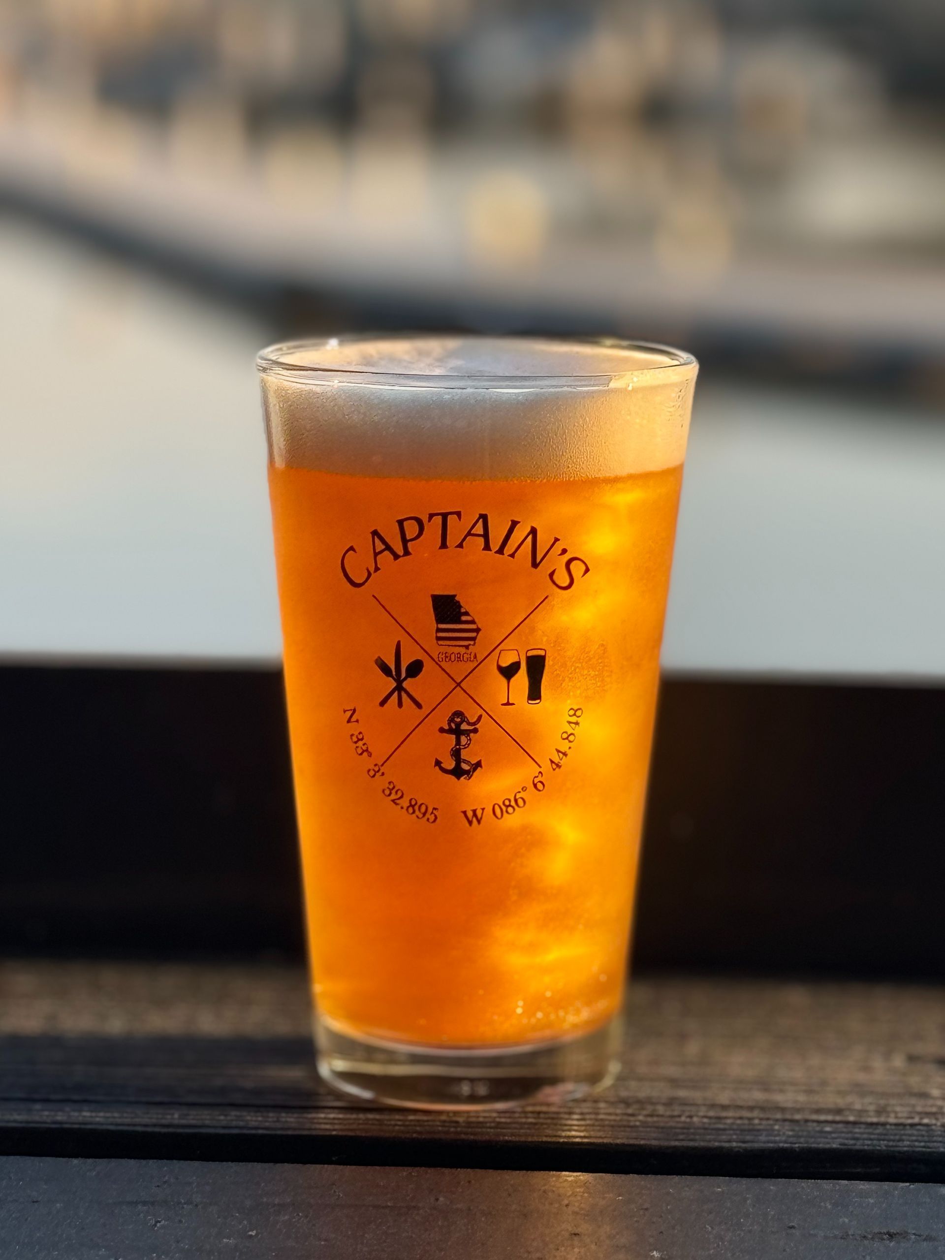 Glass of Captain's beer with foamy head, set on a dark surface, with a blurry outdoor backdrop.