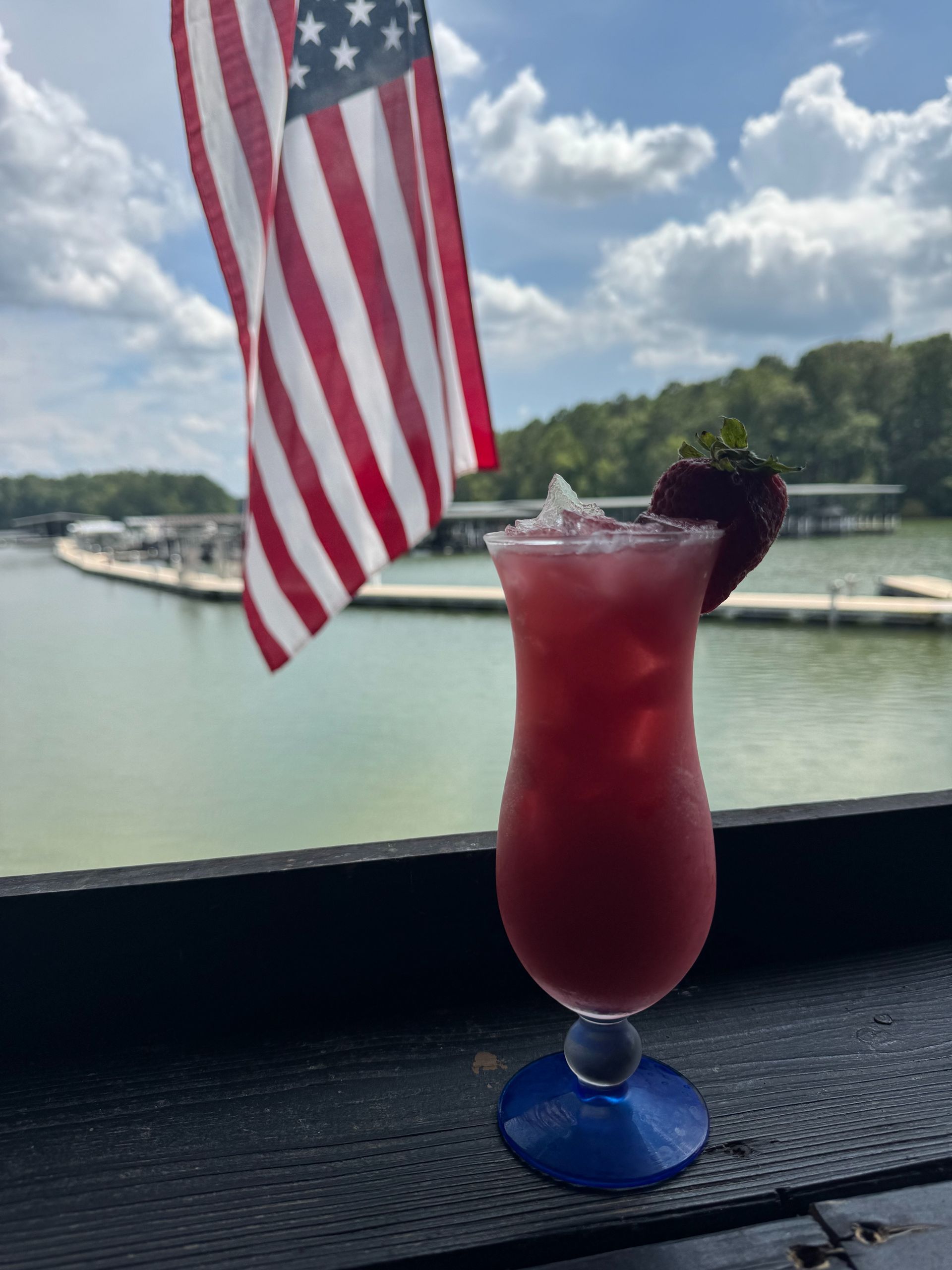 Cocktail with a strawberry garnish in front of a lake and American flag.