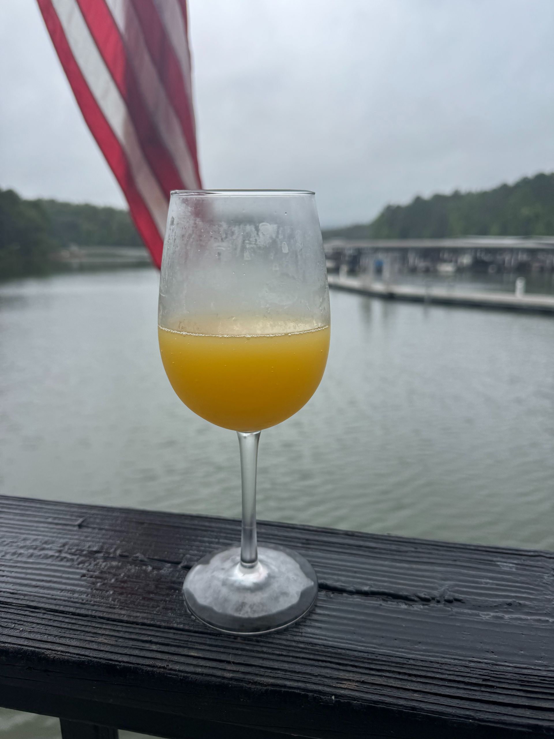 Glass of orange drink on a wooden deck, overlooking a lake with docks and an American flag.