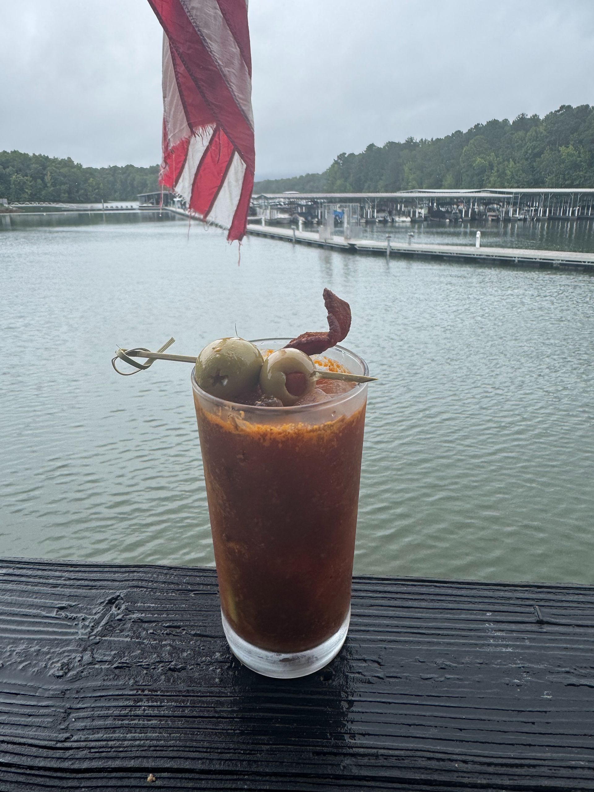 Bloody Mary cocktail on a wooden surface with a marina and American flag in the background.