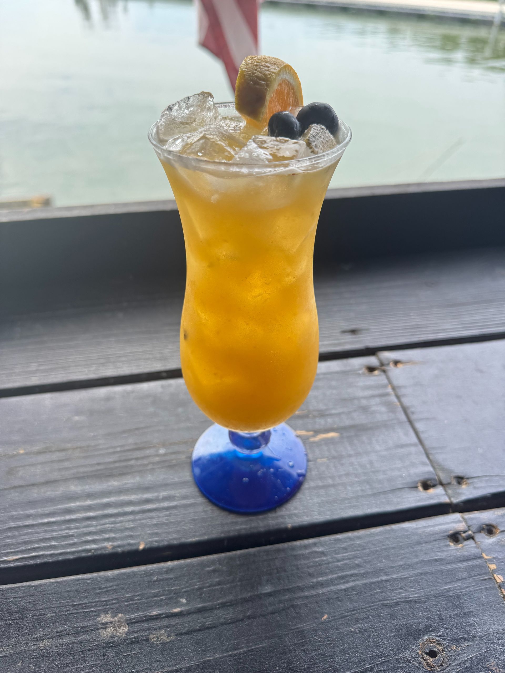 Tall yellow cocktail in a blue-based glass, garnished with ice, a small fruit, and blueberries, on a wooden surface near water.