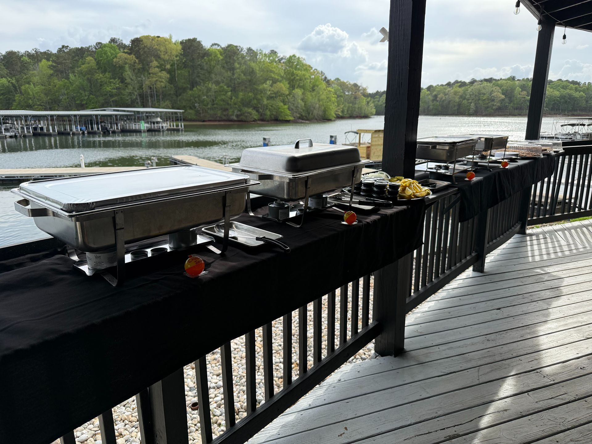 Buffet table with food on a deck overlooking a lake, stainless steel chafing dishes, black table cloths.