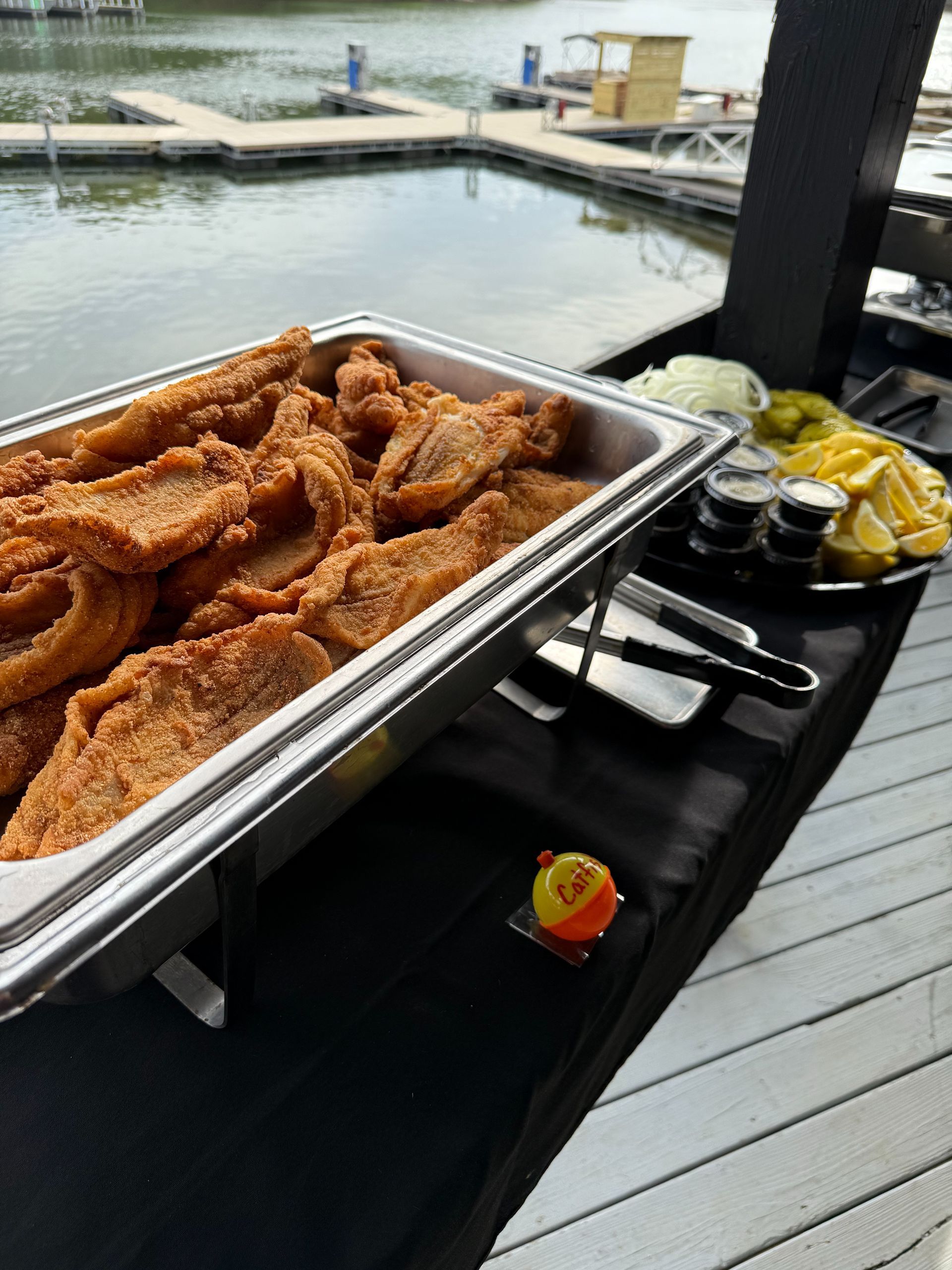 Fried fish in a buffet warmer on a table, with condiments and a waterfront in the background.