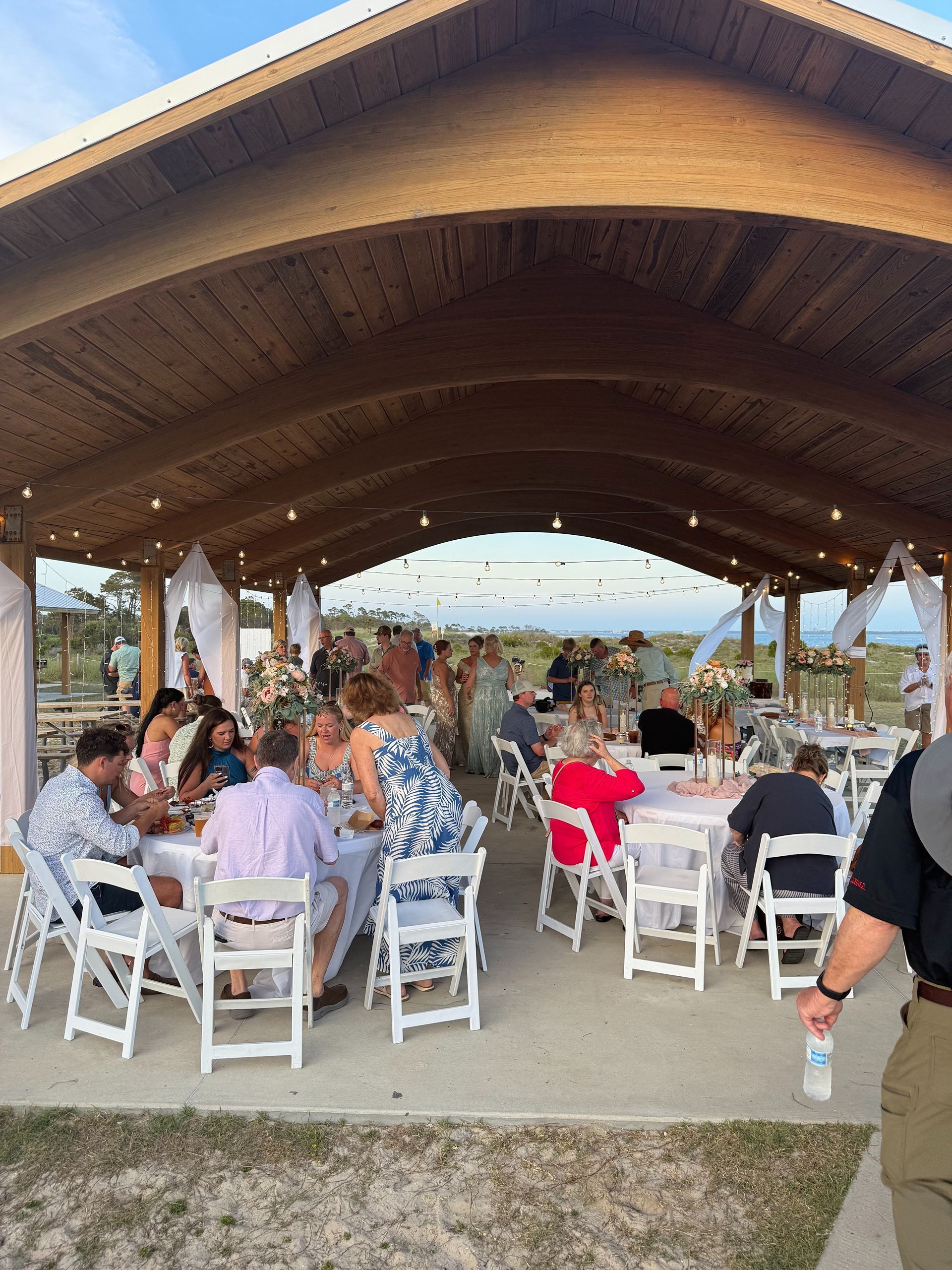 Wedding reception under a covered pavilion. Guests at round tables, some standing. Sky and water visible in background.