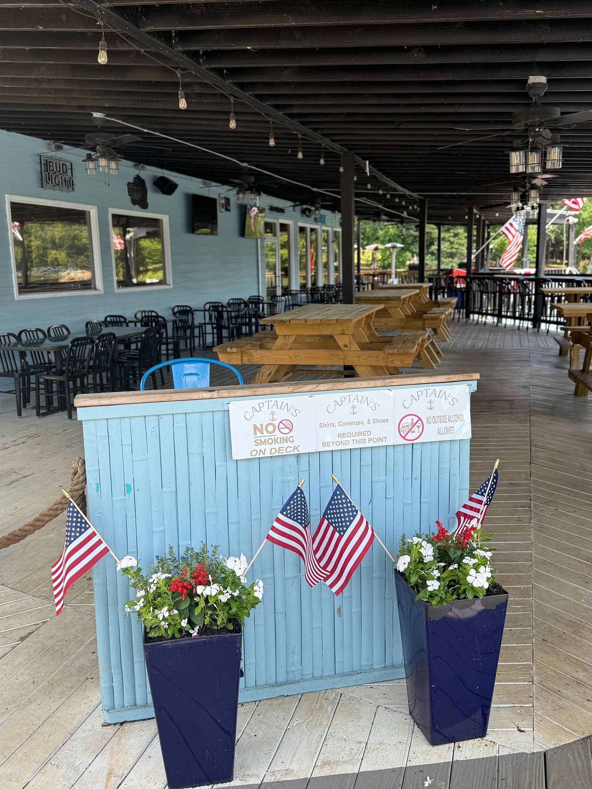 Outdoor bar with American flags and flower pots; blue facade and tables.