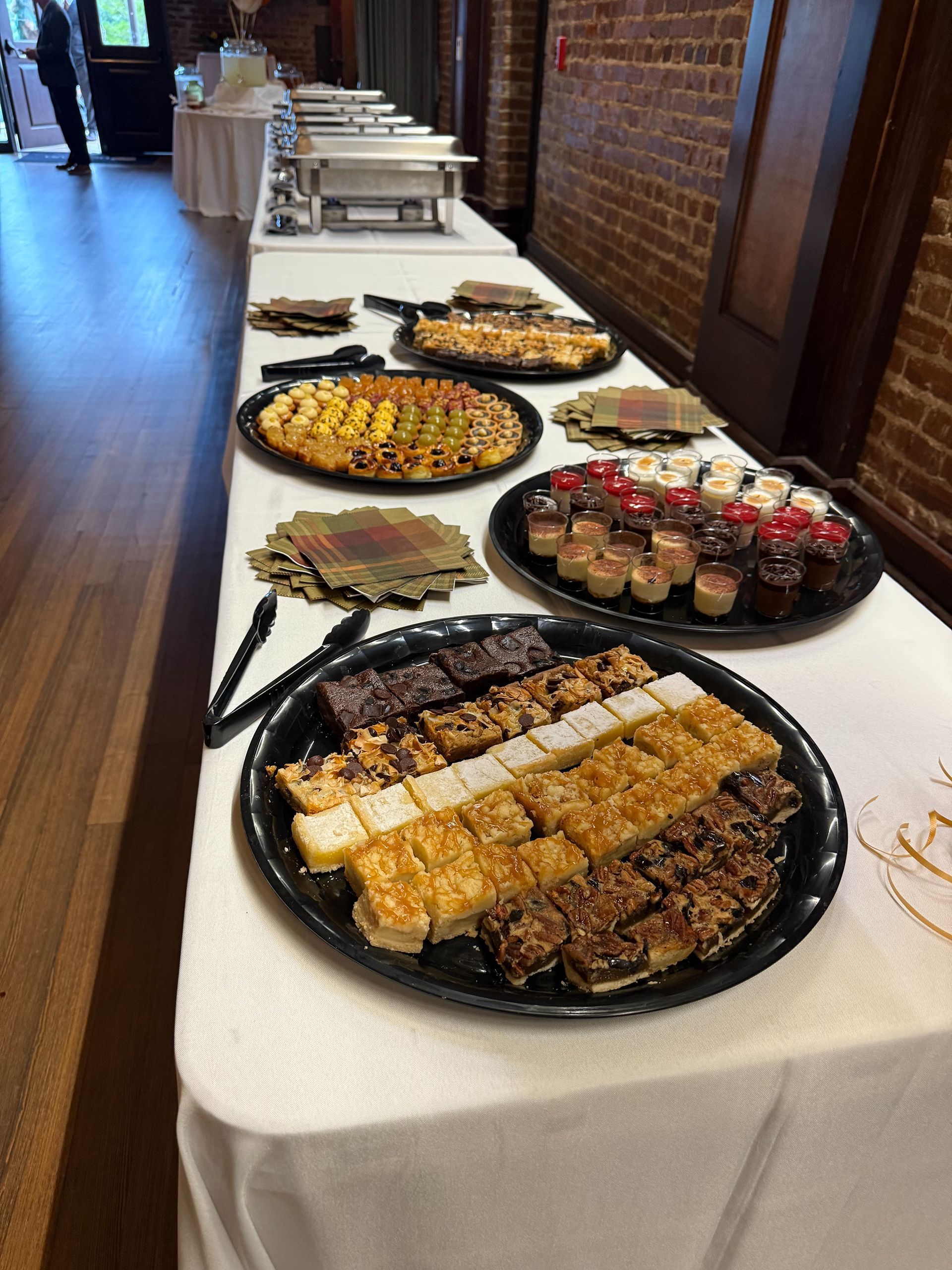 Buffet table with various desserts, trays, and chafing dishes; brick wall in background.