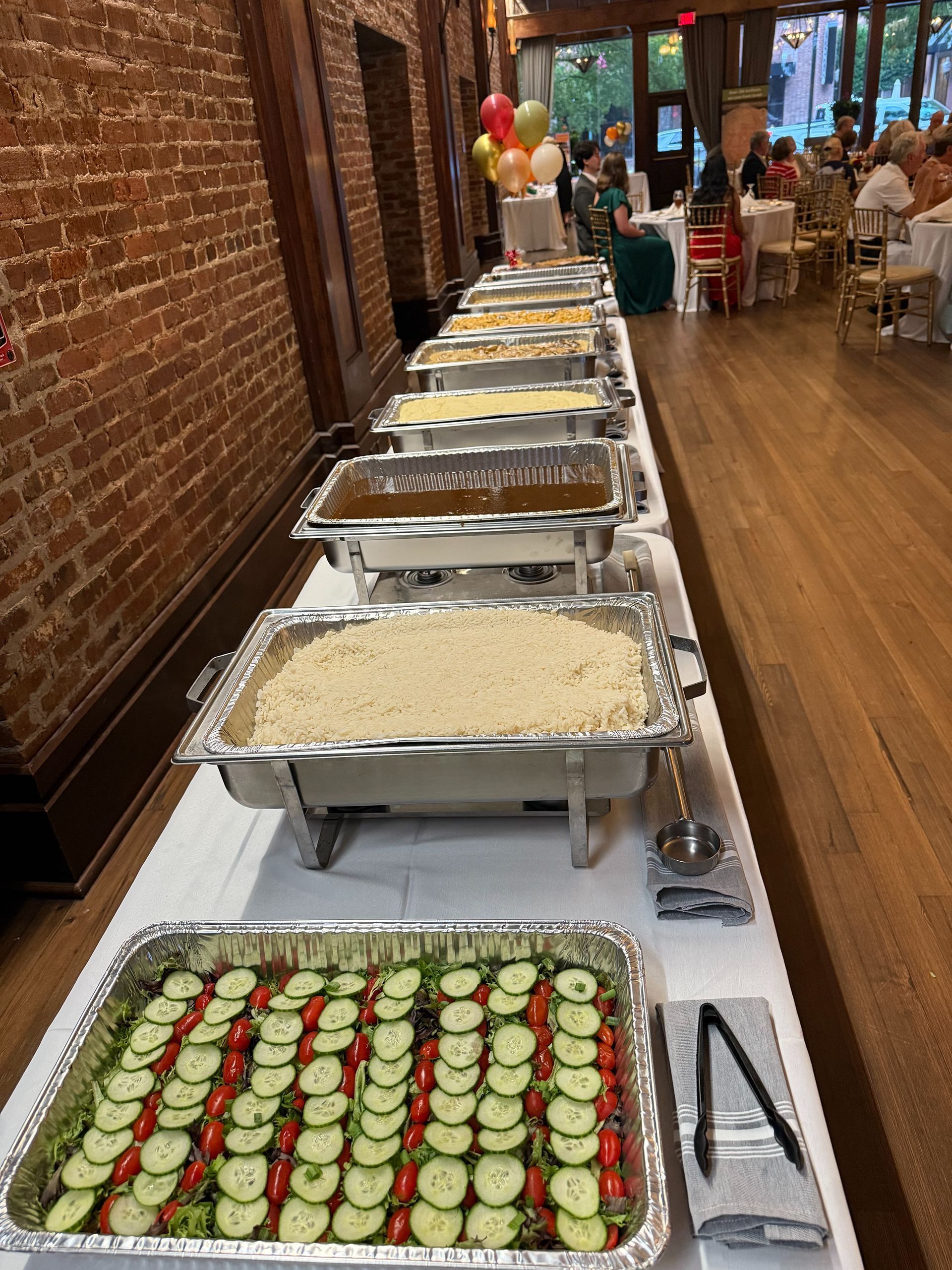 Buffet table with various dishes; a long table in a room with a brick wall and seated guests in the background.