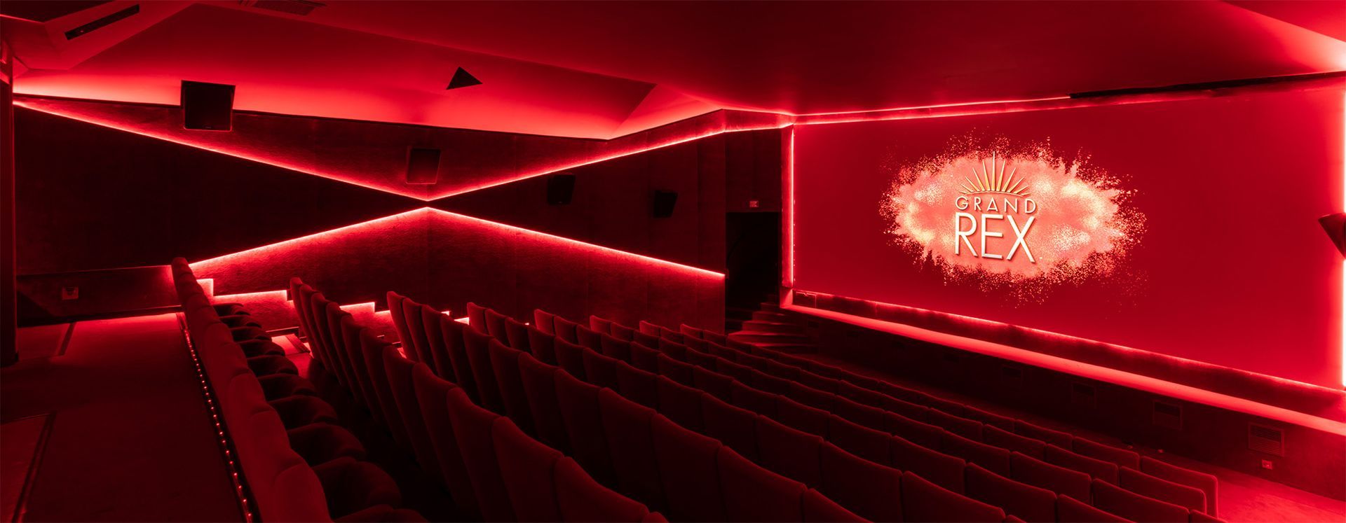 Auditorium moderne éclairé en rouge, avec des rangées de sièges et un écran abstrait lumineux sur la façade.