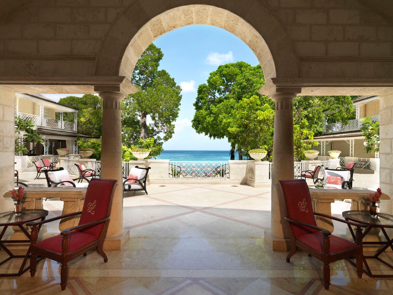 View through an archway of a patio with red chairs, toward the ocean and trees.