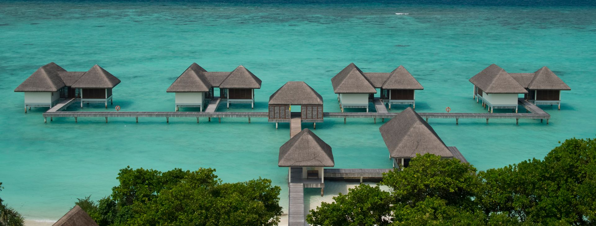 Overwater bungalows on stilts in turquoise water, under a blue sky. Lush green trees in the foreground.