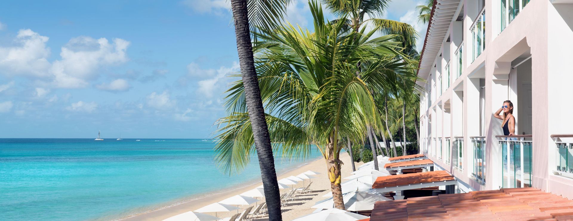 Beachfront hotel with palm trees, a sandy beach, and turquoise water. A person stands on a balcony.