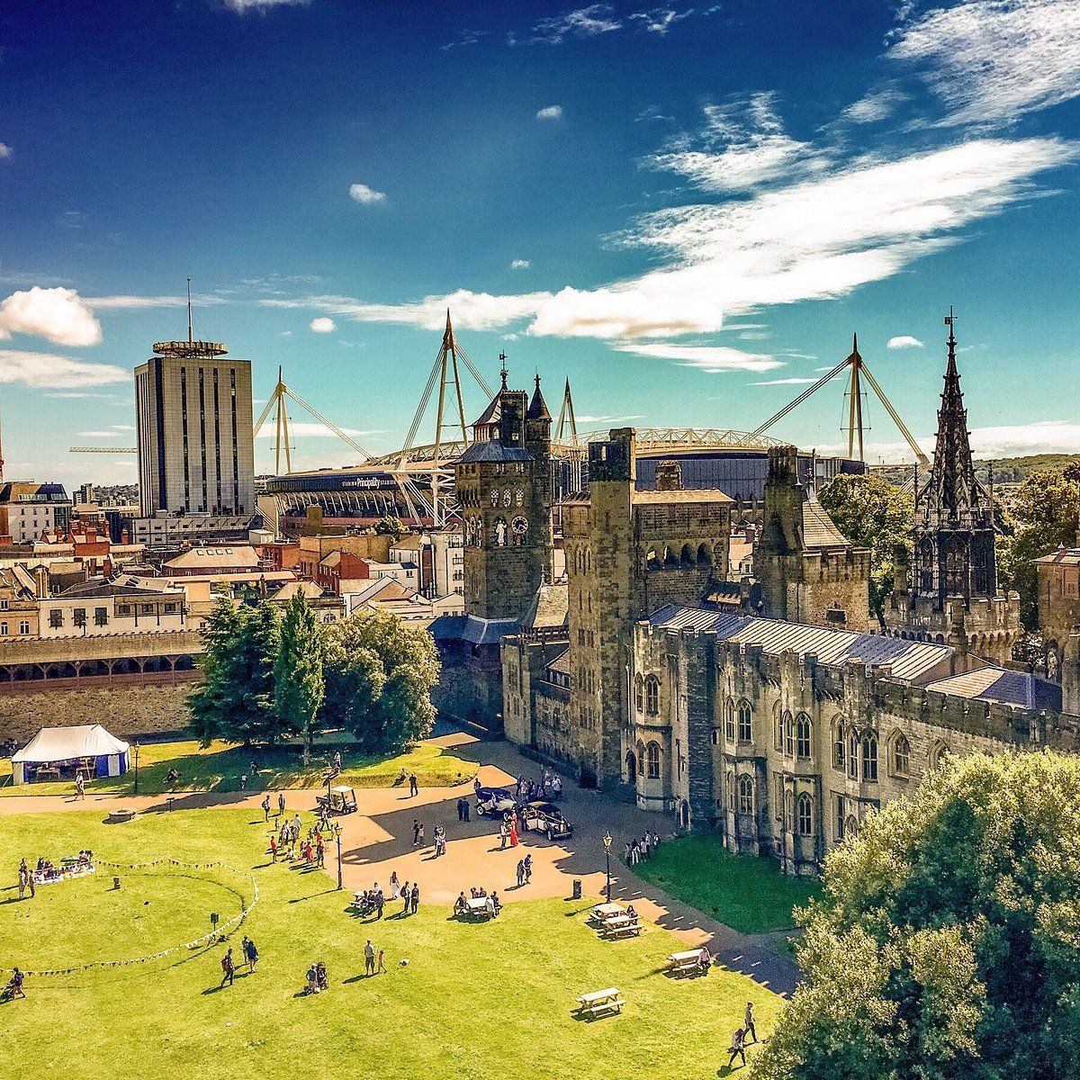 Cardiff city view: castle, green park, stadium, high-rise buildings, blue sky. People gathered.