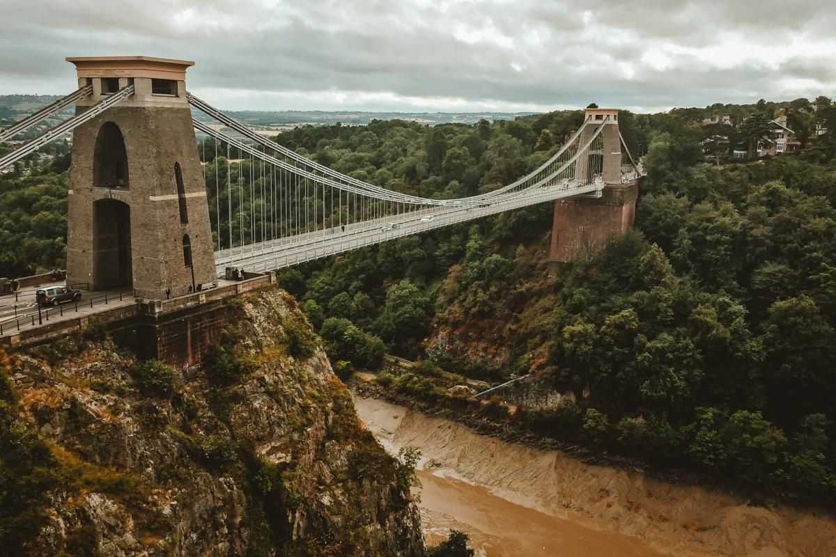 Bristol. Clifton Suspension Bridge spanning a gorge, stone towers, cables, lush green trees, overcast sky.