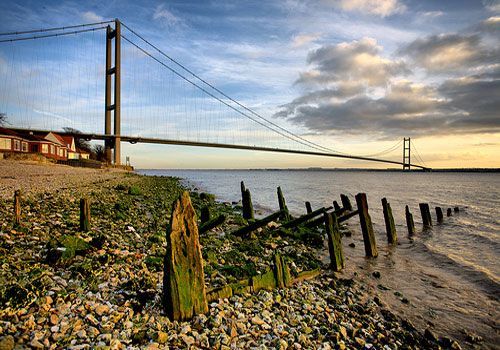 Humber Bridge over a rocky beach with weathered wooden posts, a building on the left, and a cloudy sky.