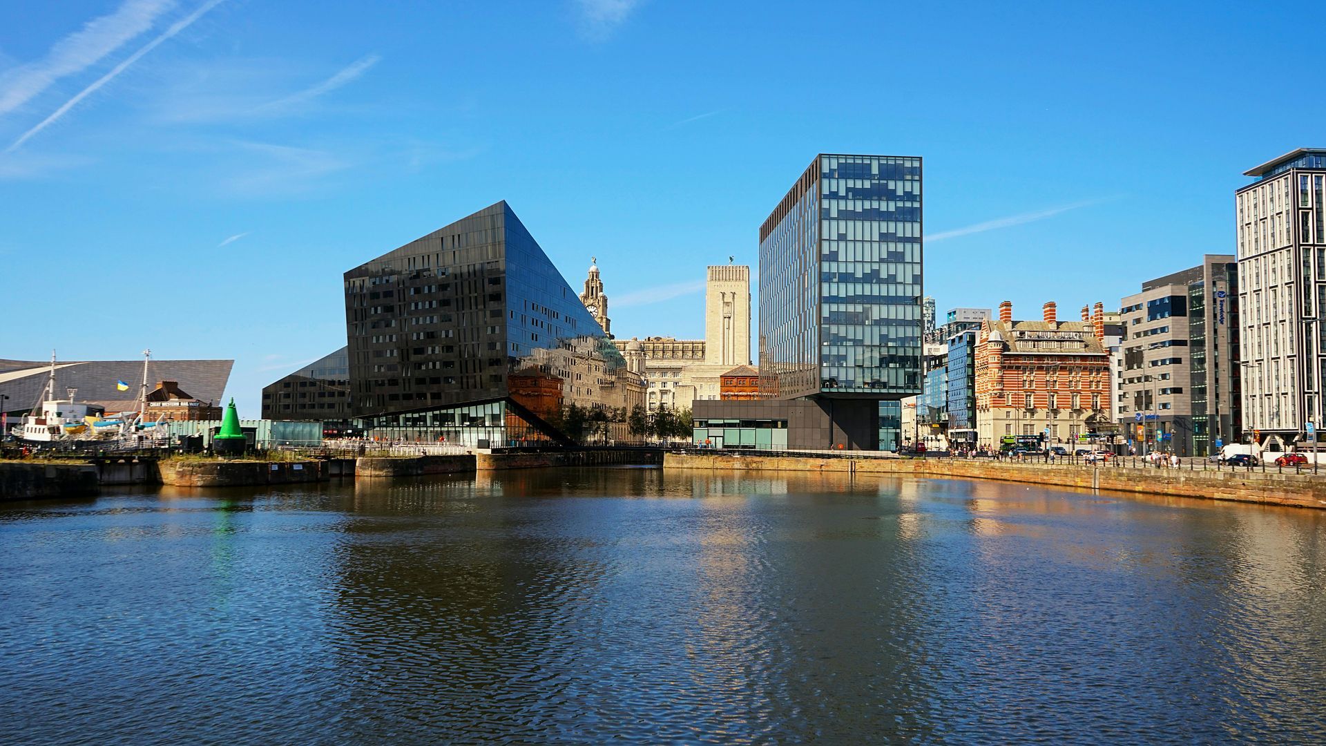 Liverpool. Waterfront buildings, including a mirrored pyramid-shaped structure, reflect on calm water under a blue sky.