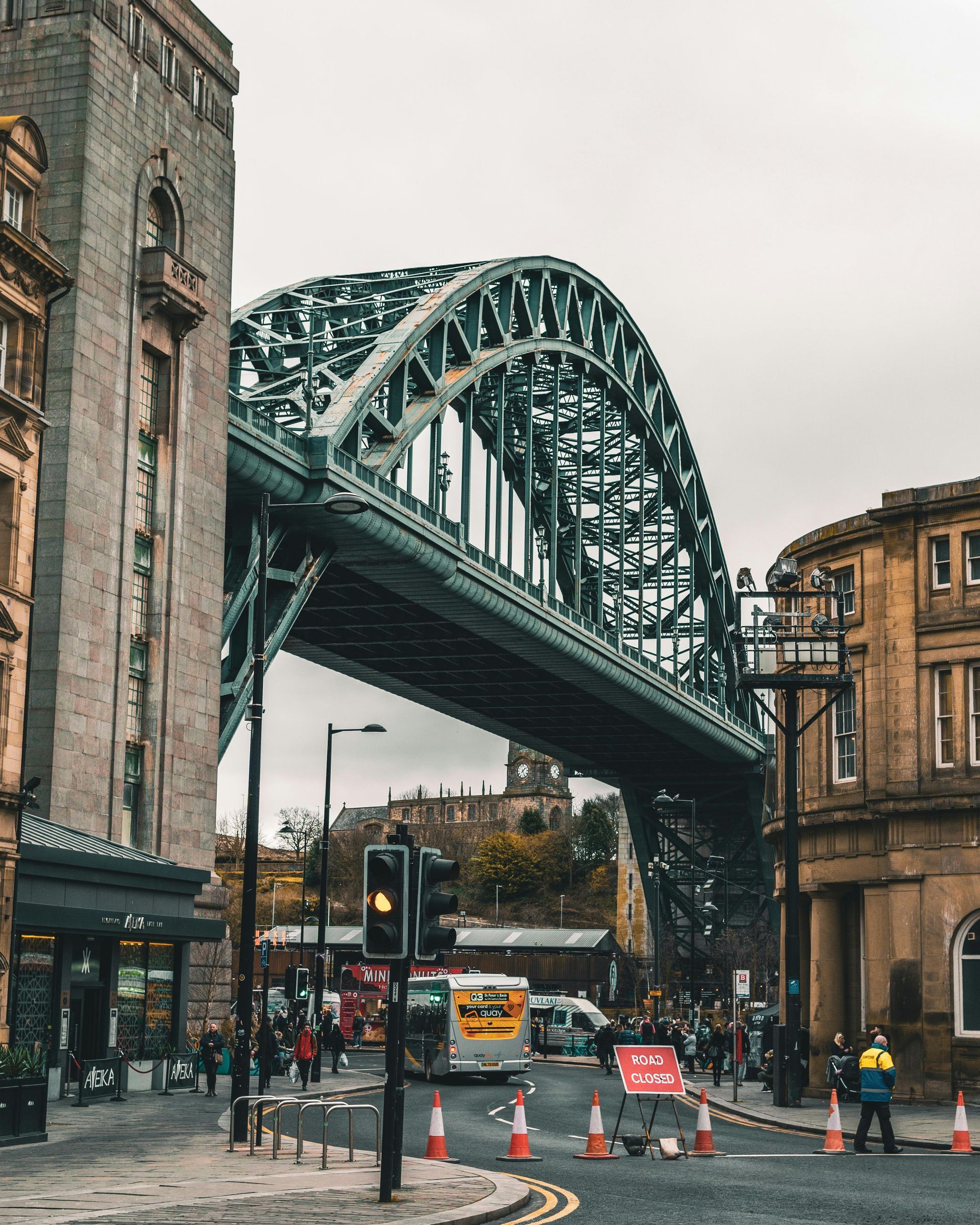 Steel arch bridge spans a street in Newcastle, UK. Buildings flank the road. Overcast sky.