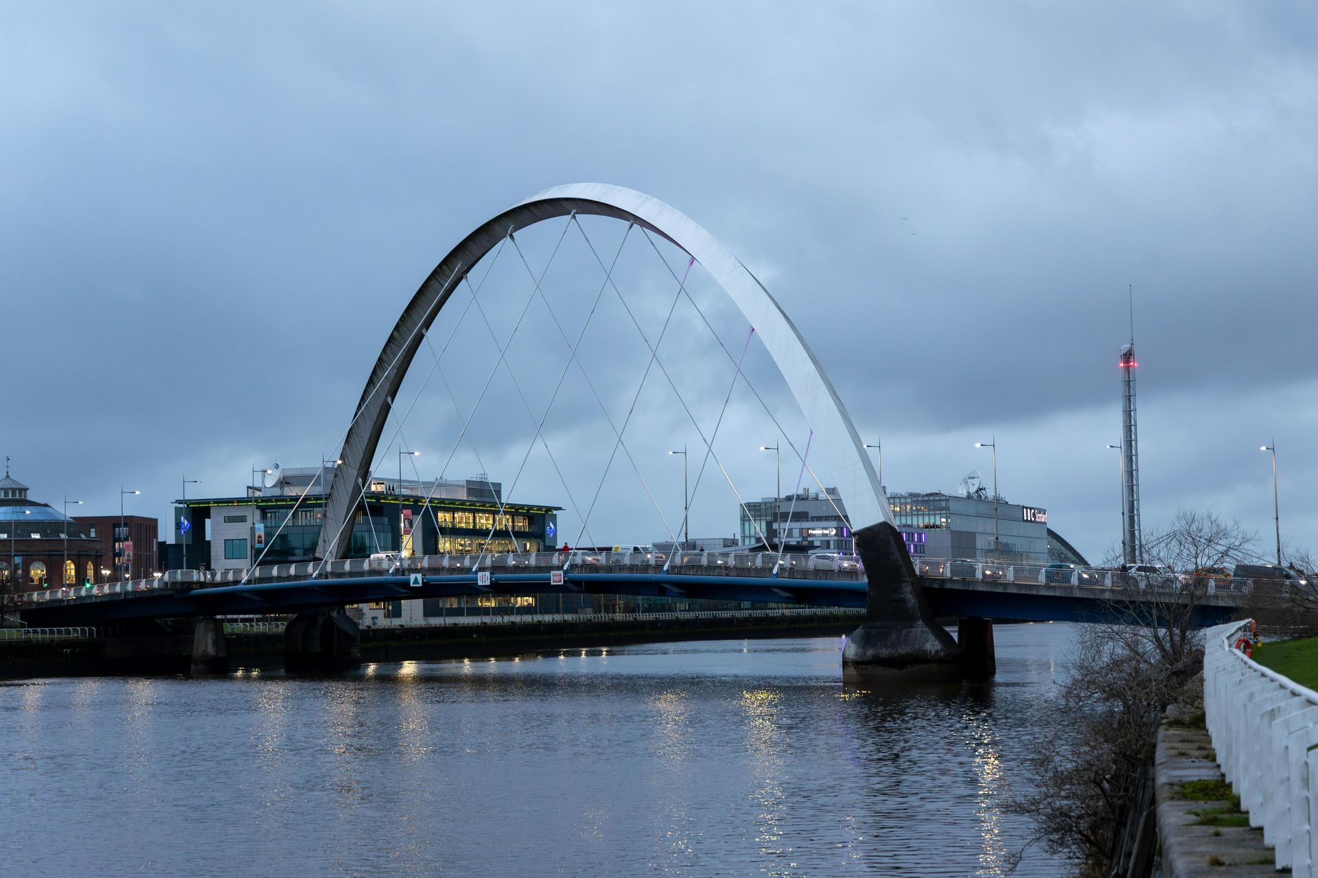 Clyde Arc bridge in Glasgow, white arch, spans the River Clyde under an overcast sky.