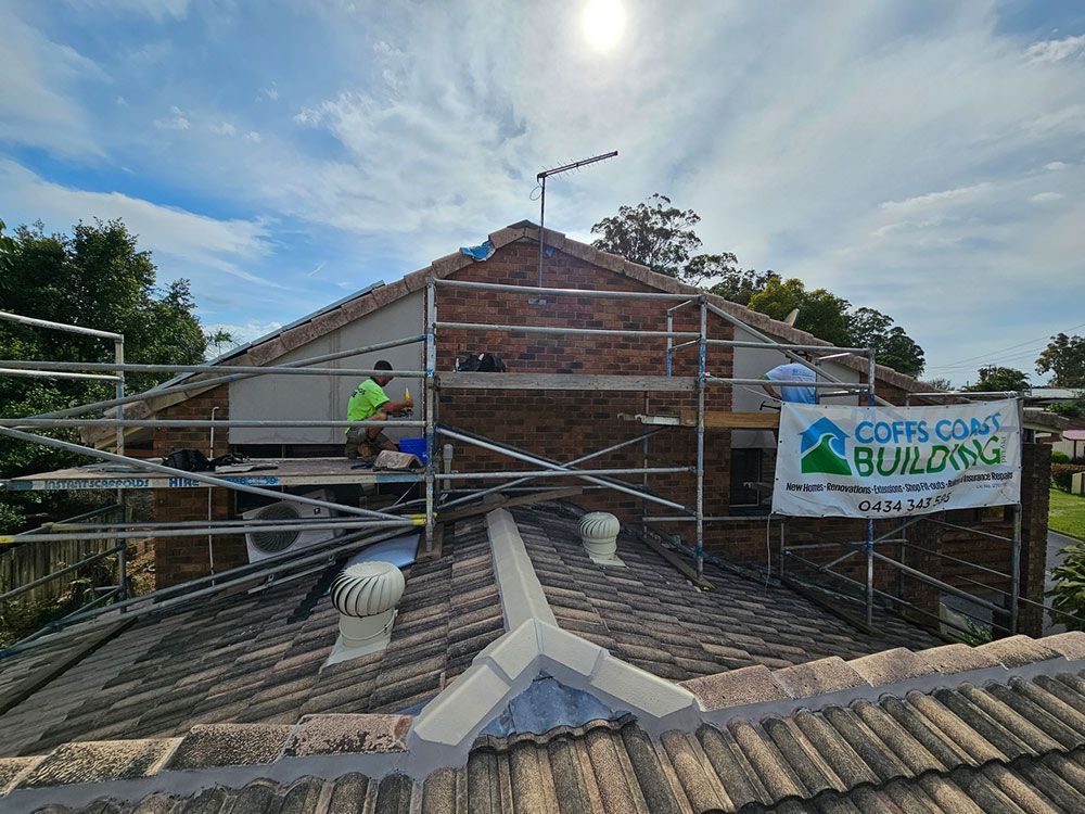 Man working on a brick wall on a house roof using scaffolding, Coffs Coast Building sign visible.