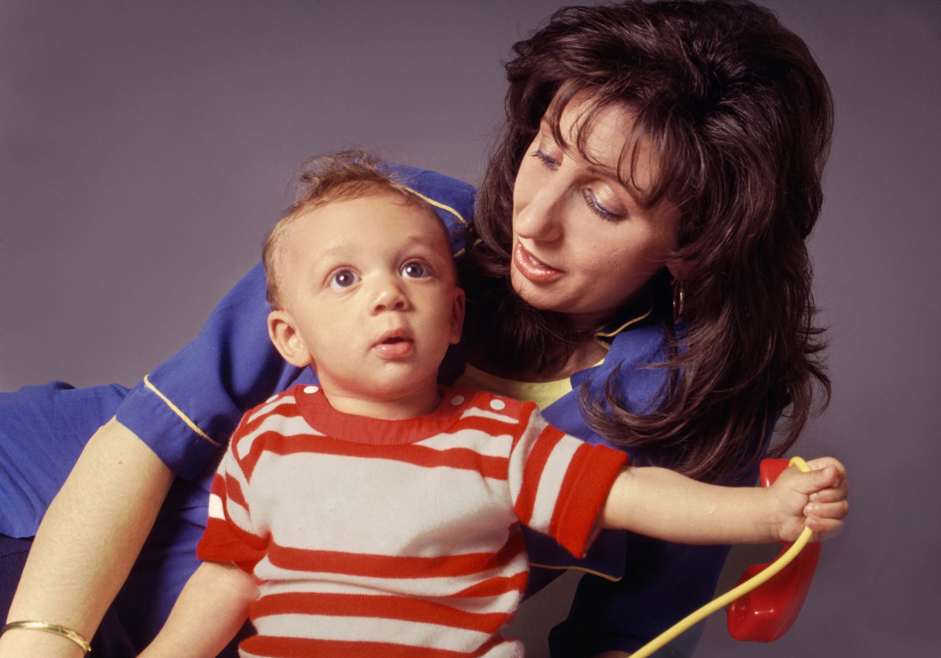 Woman leans toward a toddler holding a red toy, both looking up with interest, studio setting.