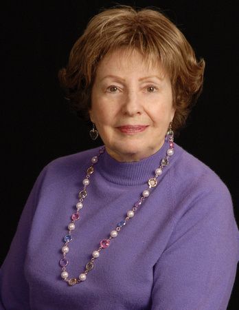 a woman in a purple sweater and pearl necklace is smiling for the camera .