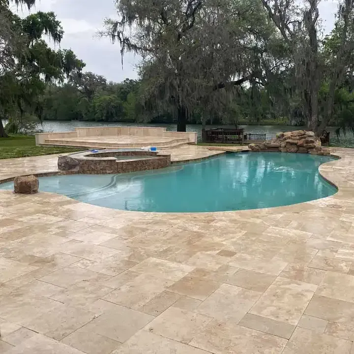 A large swimming pool is surrounded by a tiled patio next to a lake.