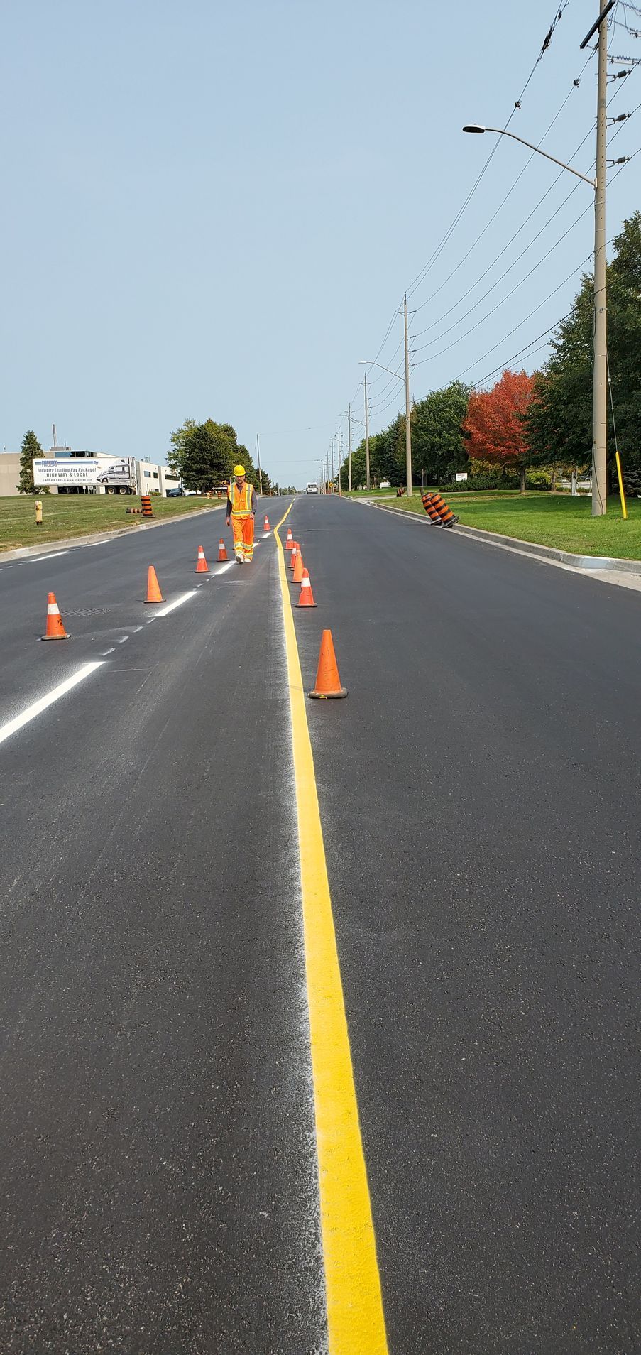 Newly paved road with orange cones and yellow center line. A worker in an orange vest stands ahead.