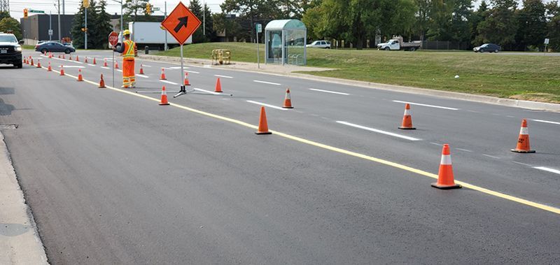 Roadwork on a freshly paved road, orange cones marking lane closures, worker in reflective gear.
