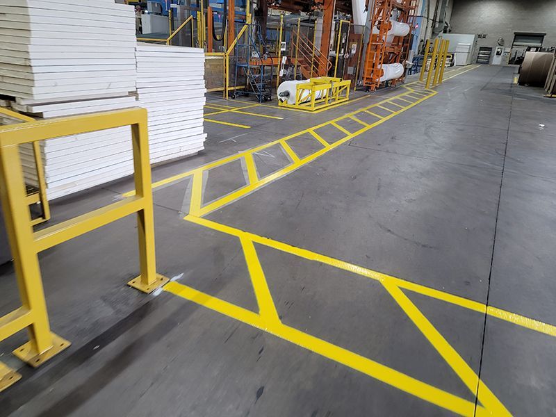 Yellow painted walkway on factory floor, next to machinery and stacks of white panels.