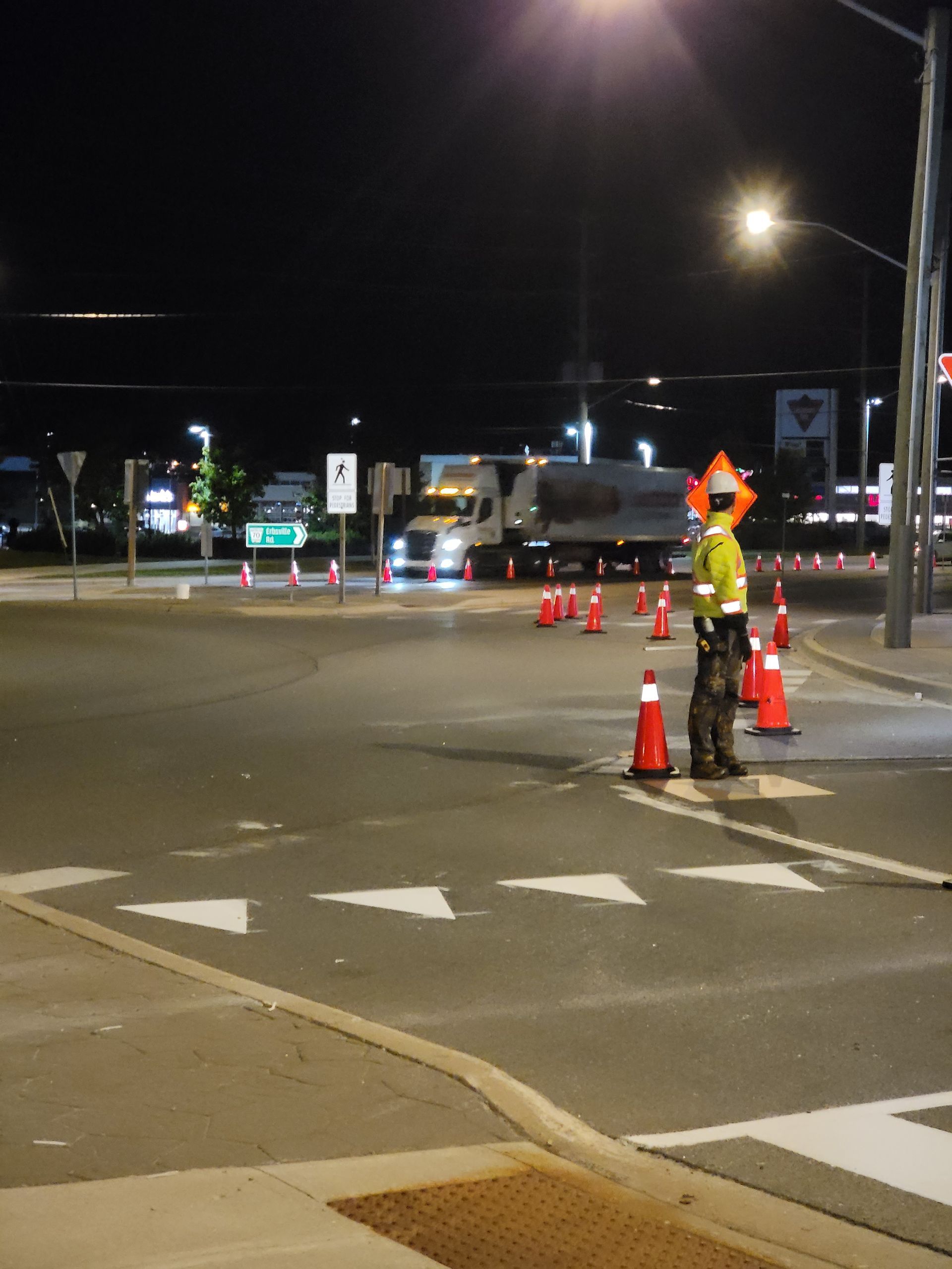 Road worker directing traffic at night, illuminated by streetlights. Cones and a semi-truck are also present.