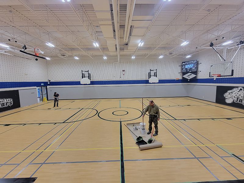 Man rolls out a rug in an empty gymnasium with another person standing in the background.
