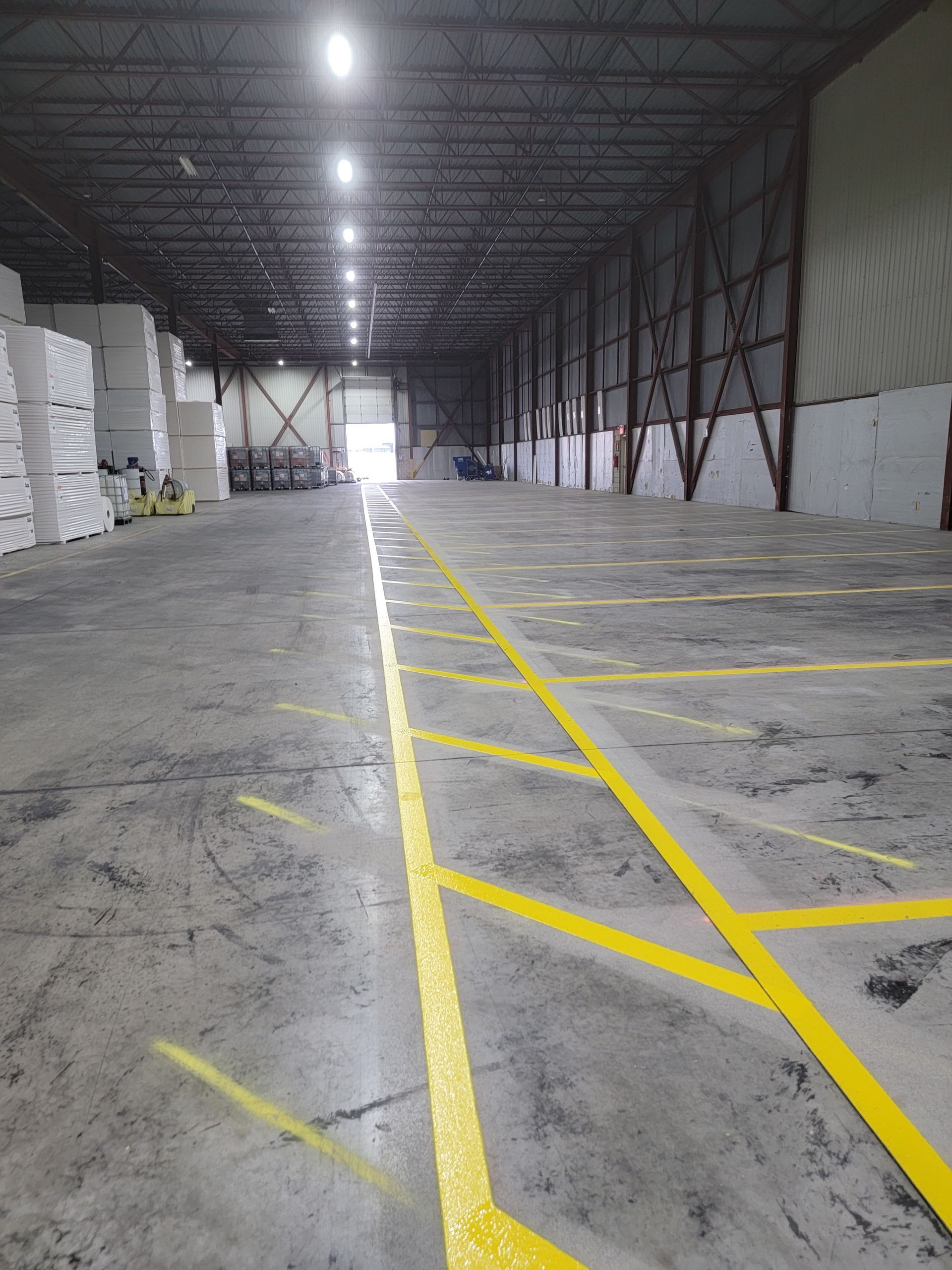 Interior of an empty warehouse with yellow floor lines. Gray concrete floor, metal structure, and bright overhead lights.