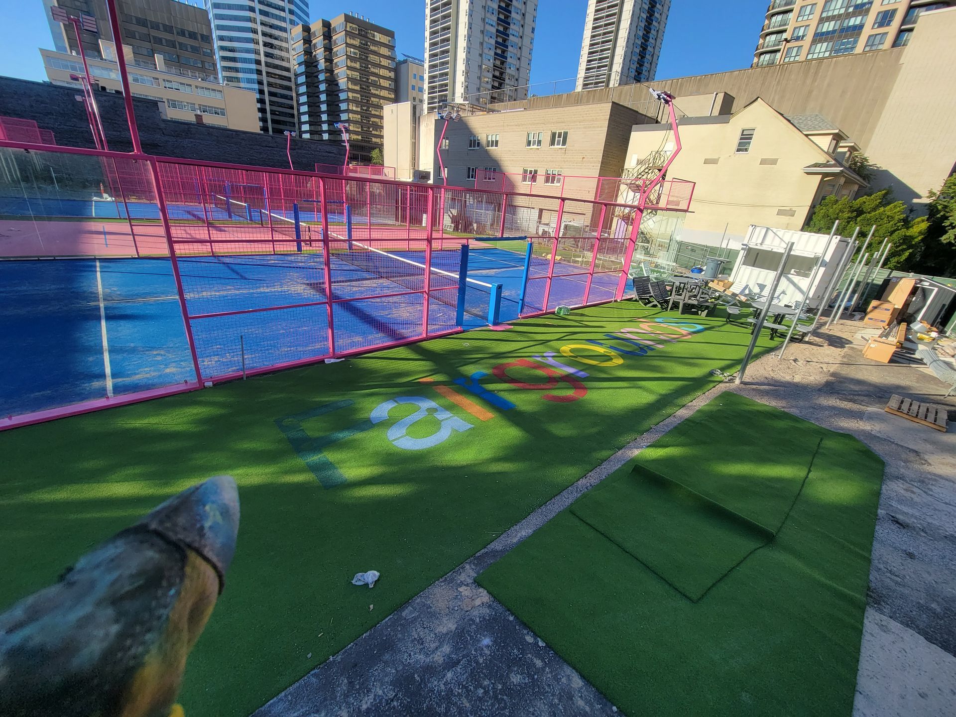 A green turf area with a blue padel court behind a pink net, with city buildings in the background.