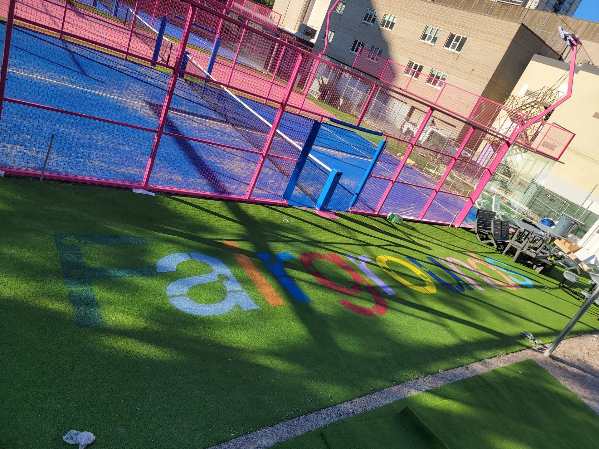Padel court with a bright pink frame and a blue playing surface. Colorful word painted on the green turf.