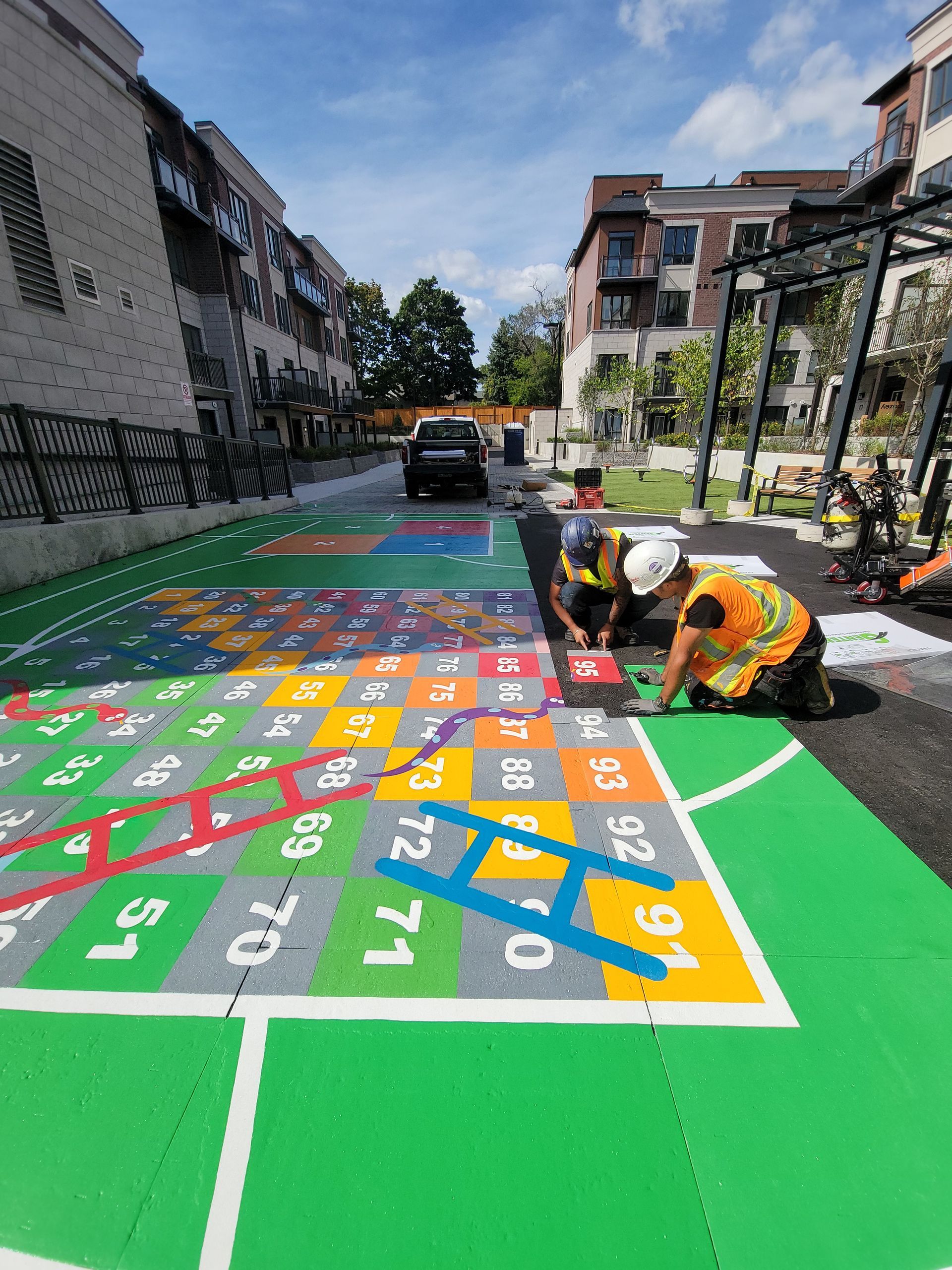 Two workers install a colorful, giant Snakes and Ladders game on a paved area. Buildings in background.