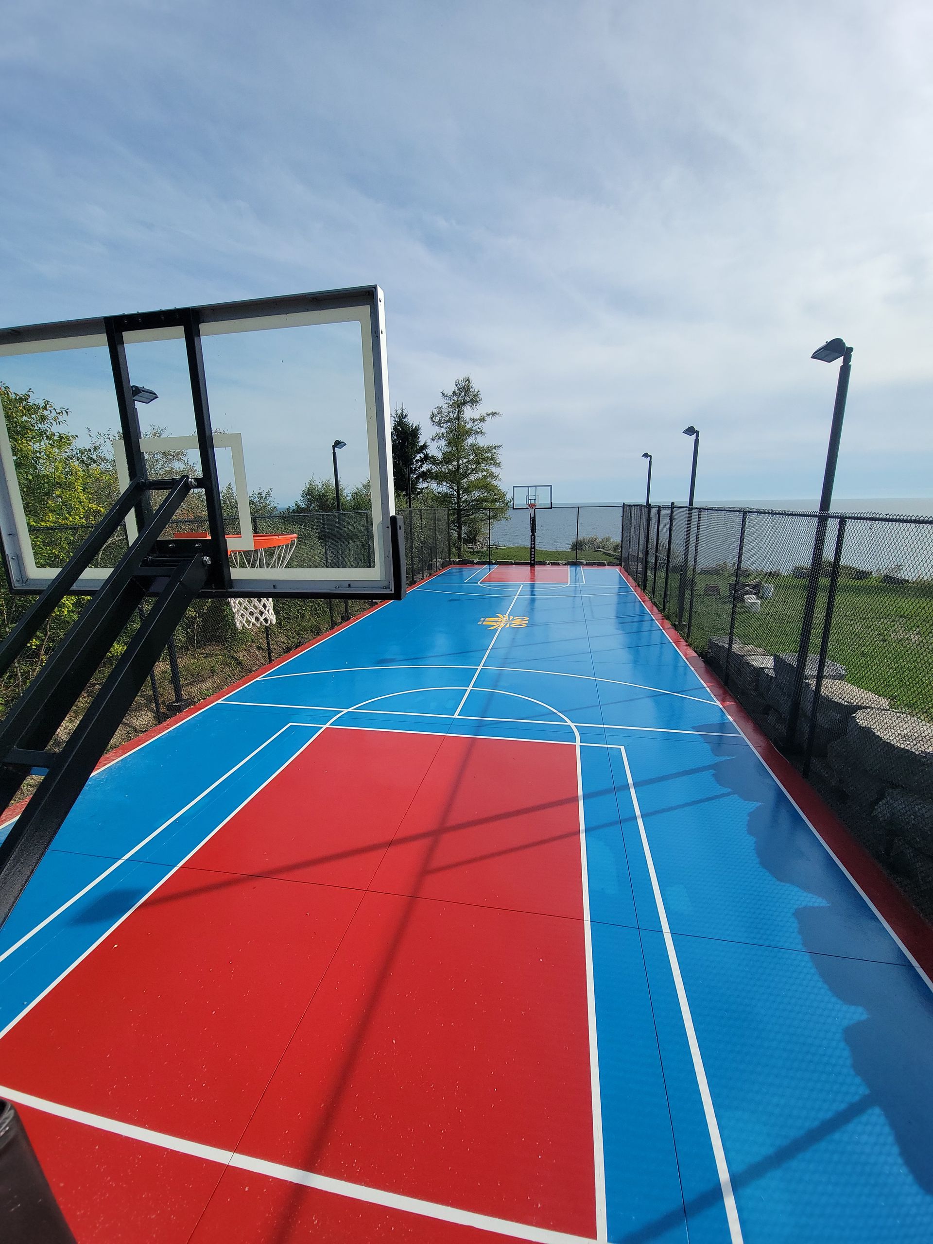 Basketball court with red and blue flooring, black net, and hoops against a cloudy sky.