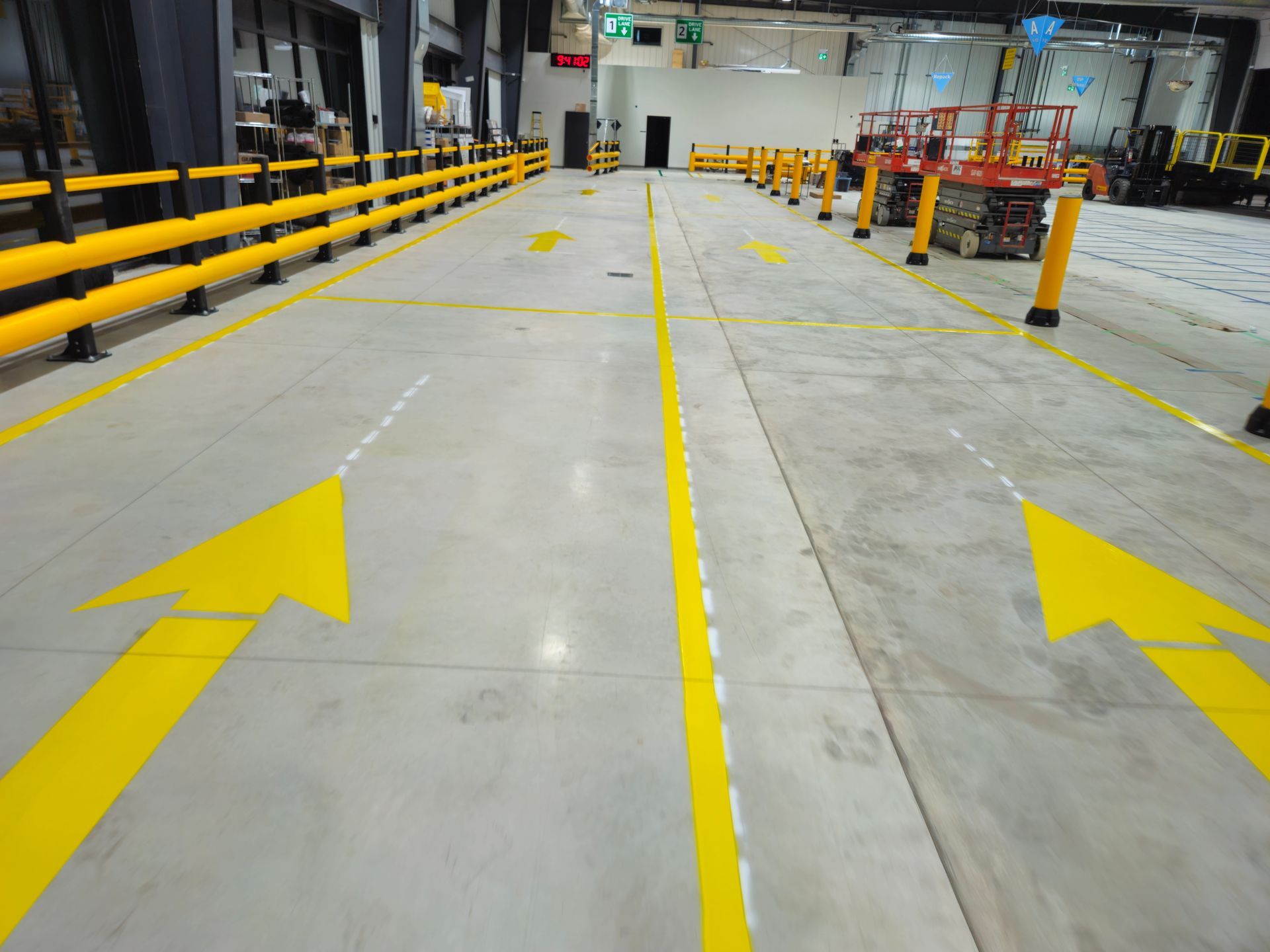 Warehouse floor with yellow arrows and lines indicating traffic flow, safety barriers visible.