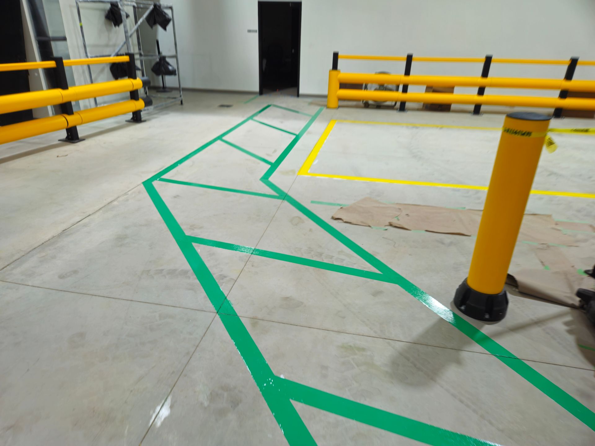 Green painted pathway on a concrete floor, with yellow barriers and bollard in an indoor space.