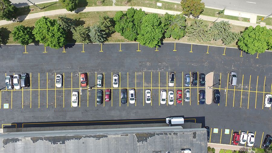 Aerial view of a parking lot with cars parked in marked spaces. Green trees and a building are adjacent.