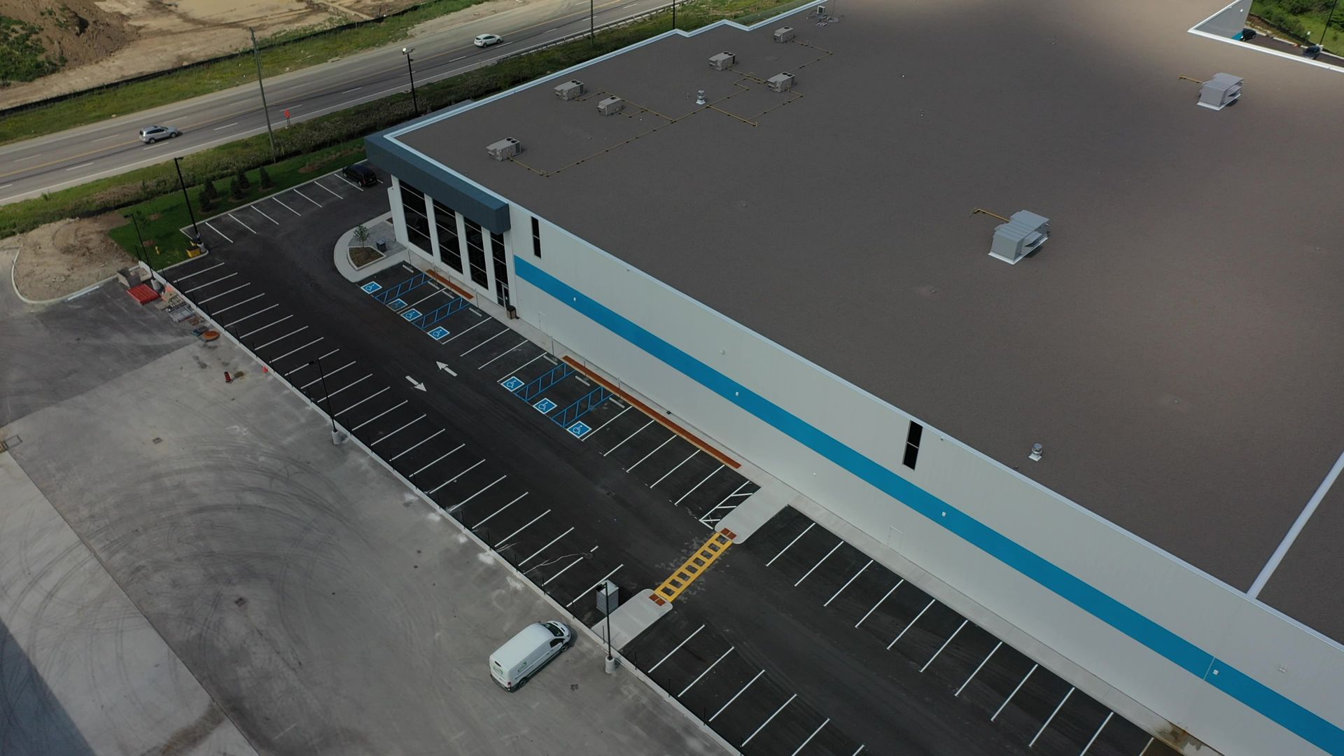 Aerial view of an Amazon warehouse with parking lot. Features include a gray roof, blue stripe, and delivery doors.