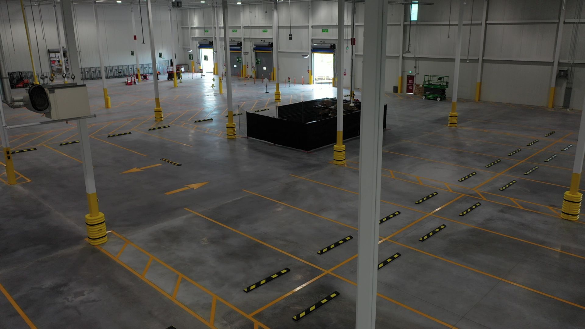 Large empty warehouse, concrete floor with yellow markings and pillars, loading doors in background.