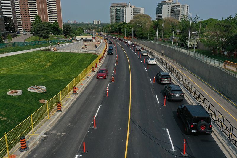 Cars driving on a multi-lane road separated by orange cones, buildings and green space in the background.