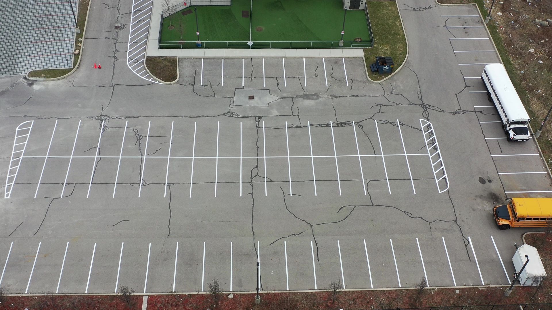 Overhead view of a cracked asphalt parking lot with white parking space lines; a bus and a school bus are parked.